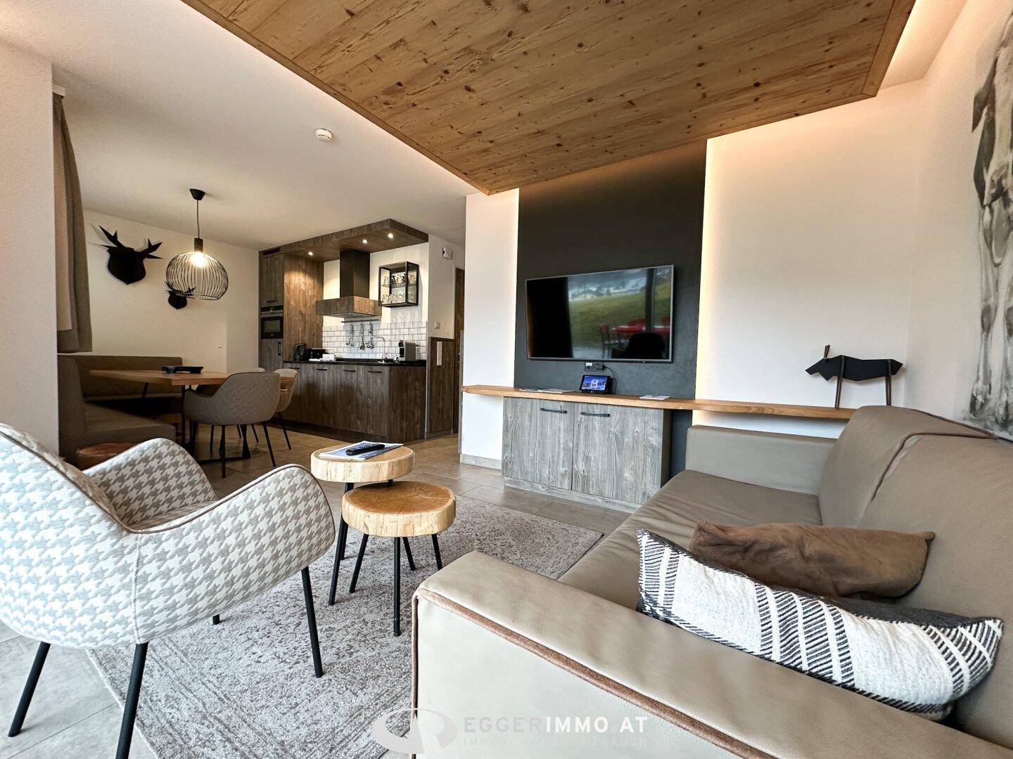 Offener Wohnbereich mit bequemer Couch, Fernseher und integrierter Küche im Hintergrund.