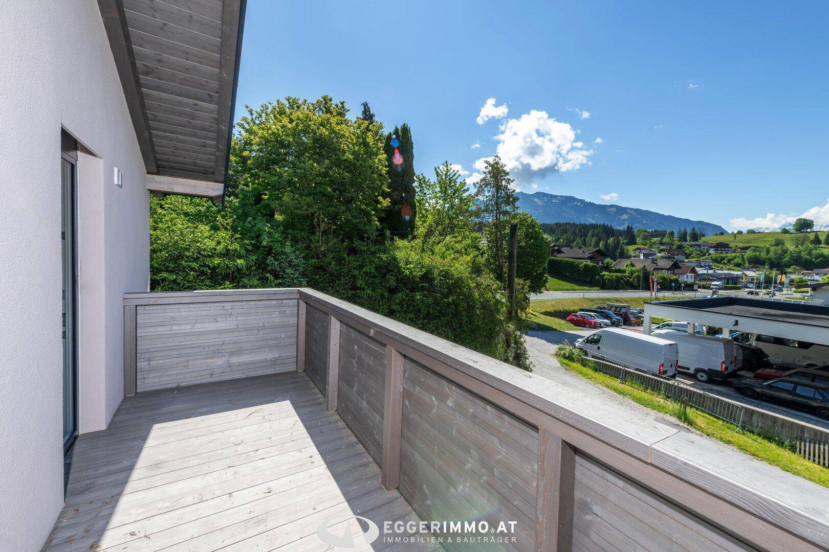 Balkon mit Holzboden und Blick auf die Straße sowie umliegende Bäume und Gebäude.