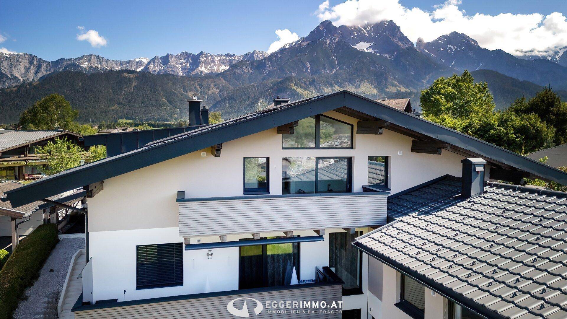 Moderne Hausfassade mit großen Fenstern und Balkon vor beeindruckender Bergkulisse.