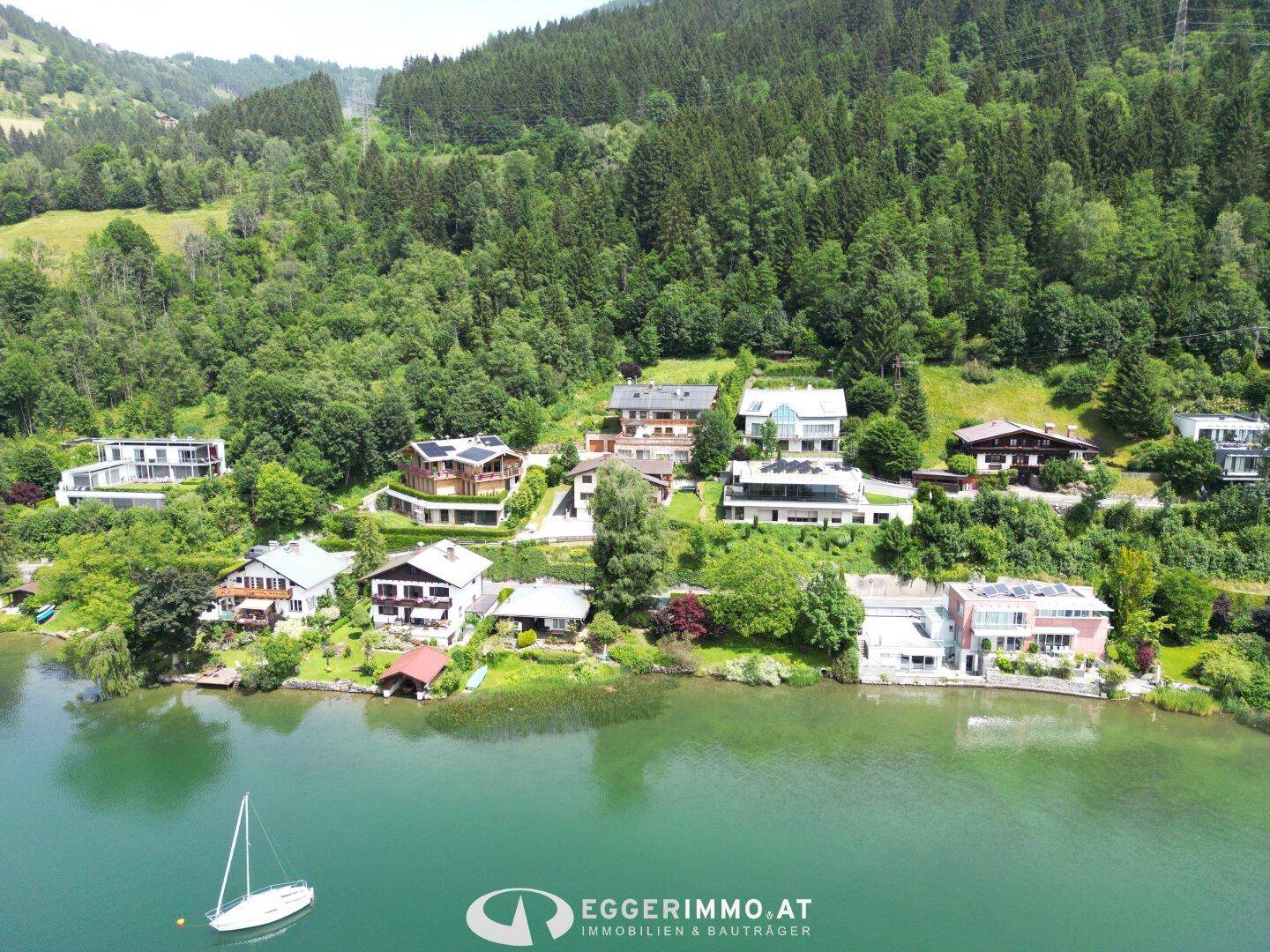 Luftaufnahme von Häusern am Seeufer mit umliegenden grünen Hügeln und einem Segelboot.