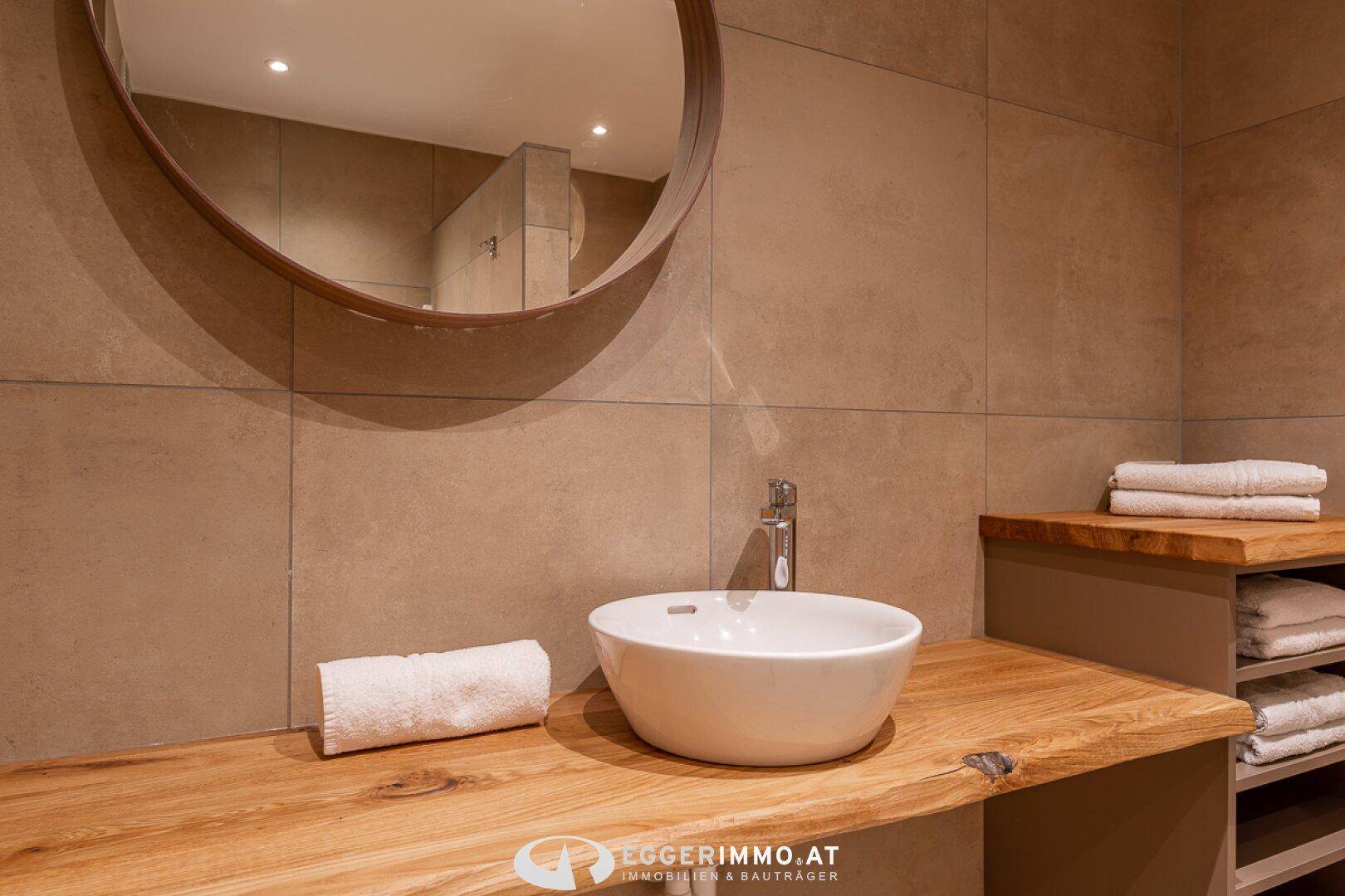 Modernes Badezimmer mit rundem Spiegel, Holzwaschtisch und beigen Fliesen.