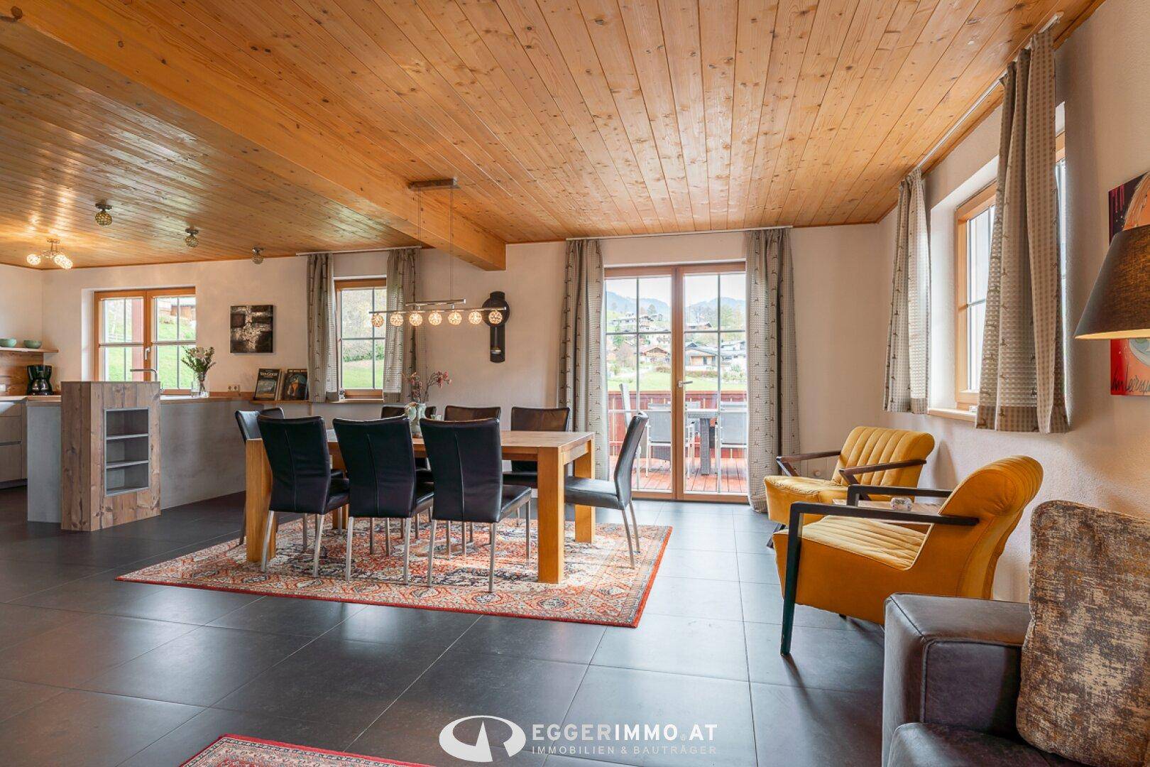 Geräumiger Wohn- und Essbereich mit Holzbalkendecke, dunklem Fliesenboden und Zugang zum Balkon mit Bergblick.