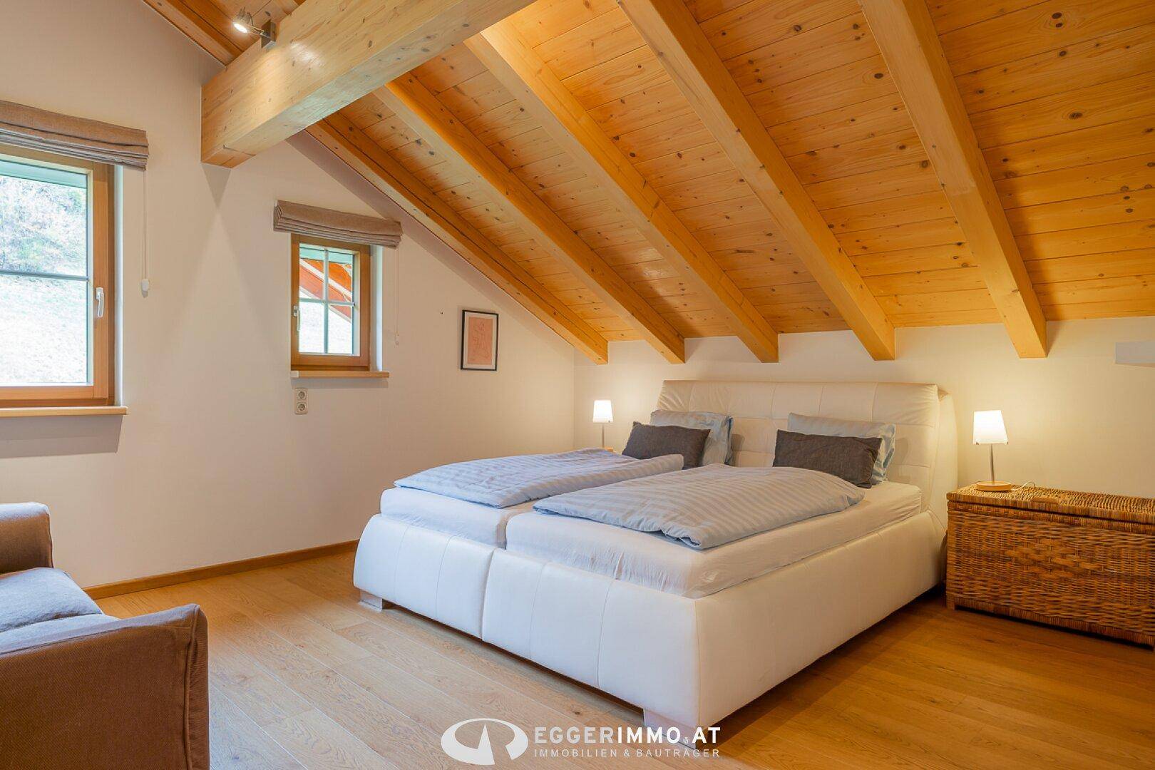 Gemütliches Schlafzimmer mit Doppelbett, Holzbalkendecke und Holzboden, hell durch zwei Fenster.