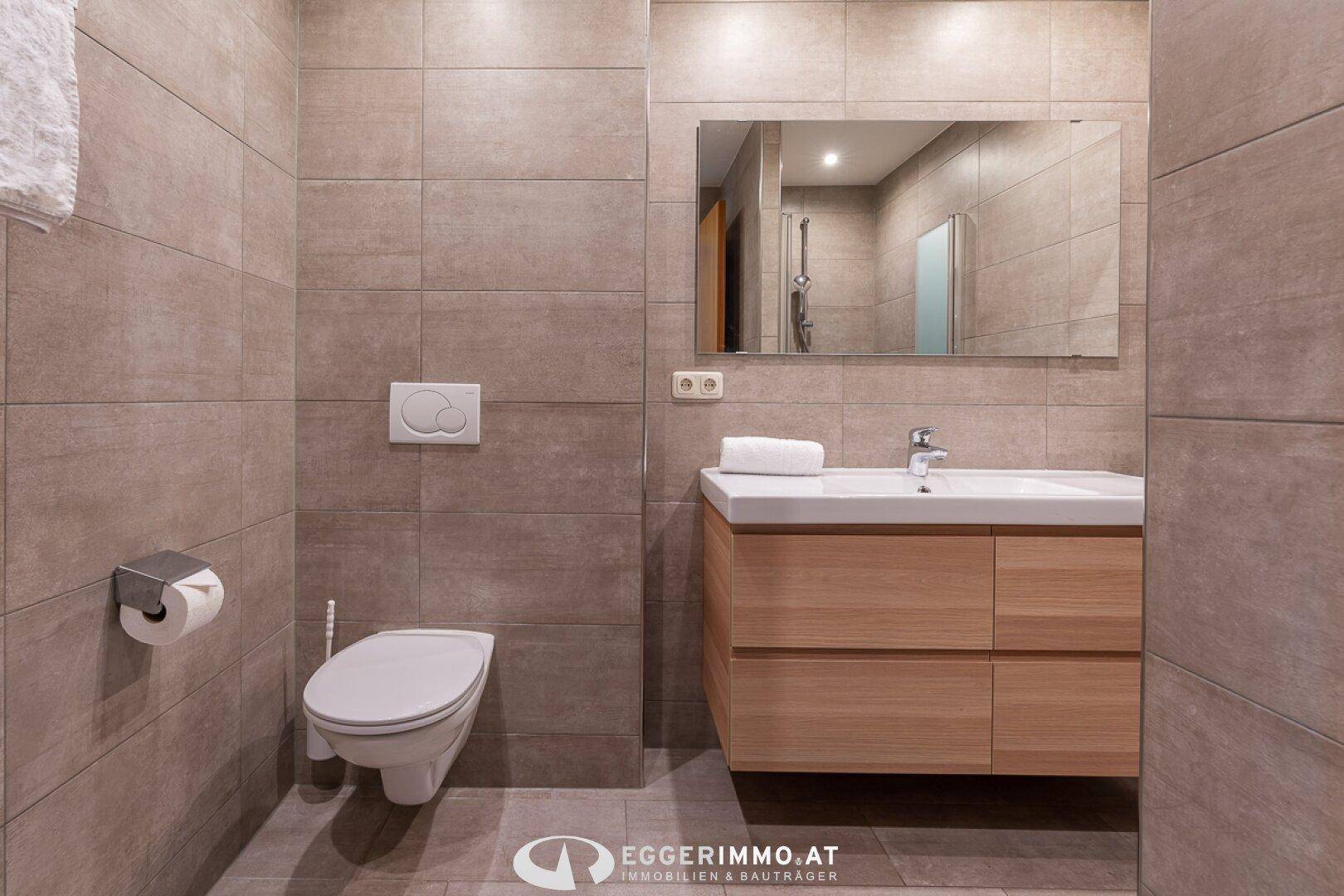 Modernes Badezimmer mit wandhängendem WC, Einzelwaschbecken und großen grauen Fliesen an Wänden und Boden.