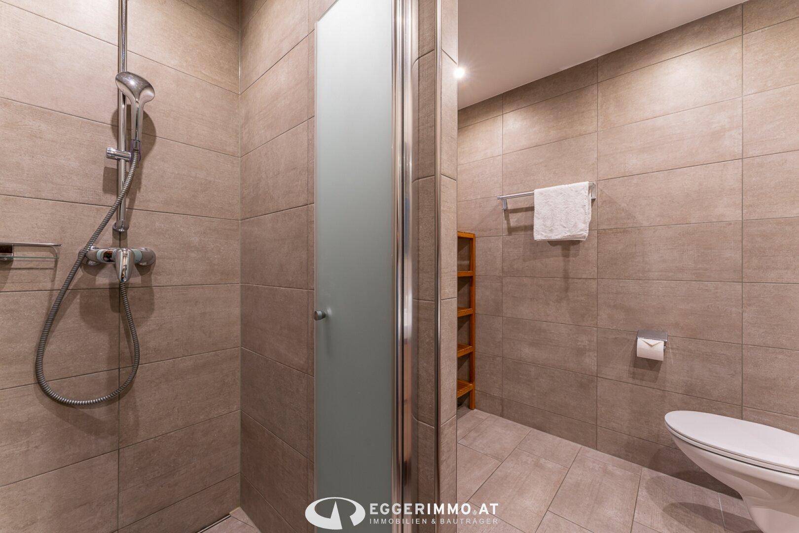 Duschbereich im Badezimmer mit grauen Fliesen, Glasduschwand und Handtuchhalter.