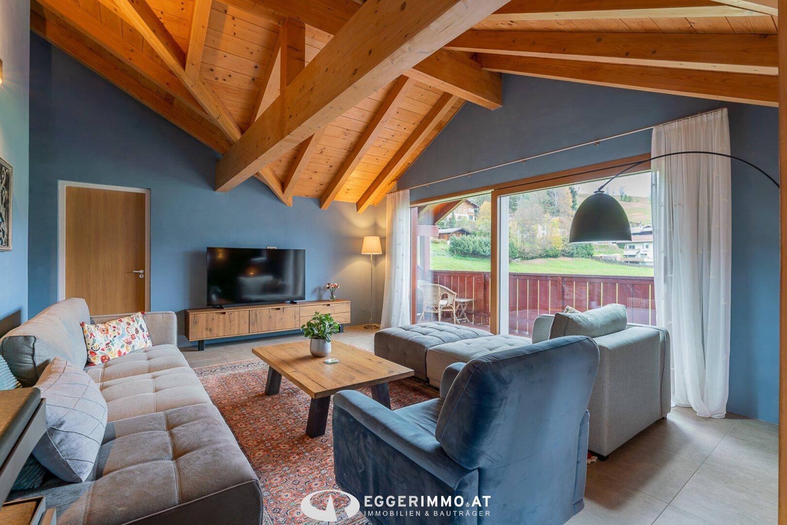 Gemütliches Wohnzimmer mit Sofa, Fernseher und großem Fenster mit Zugang zum Balkon und Blick ins Grüne.