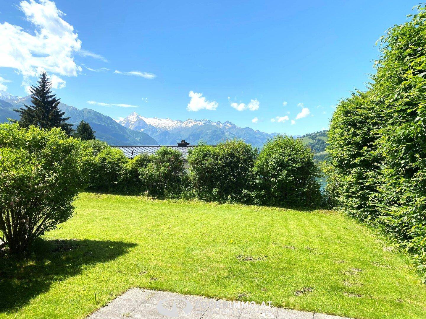 Sonniger Gartenbereich mit Rasen, Terrasse und Panoramablick auf Berge und See.