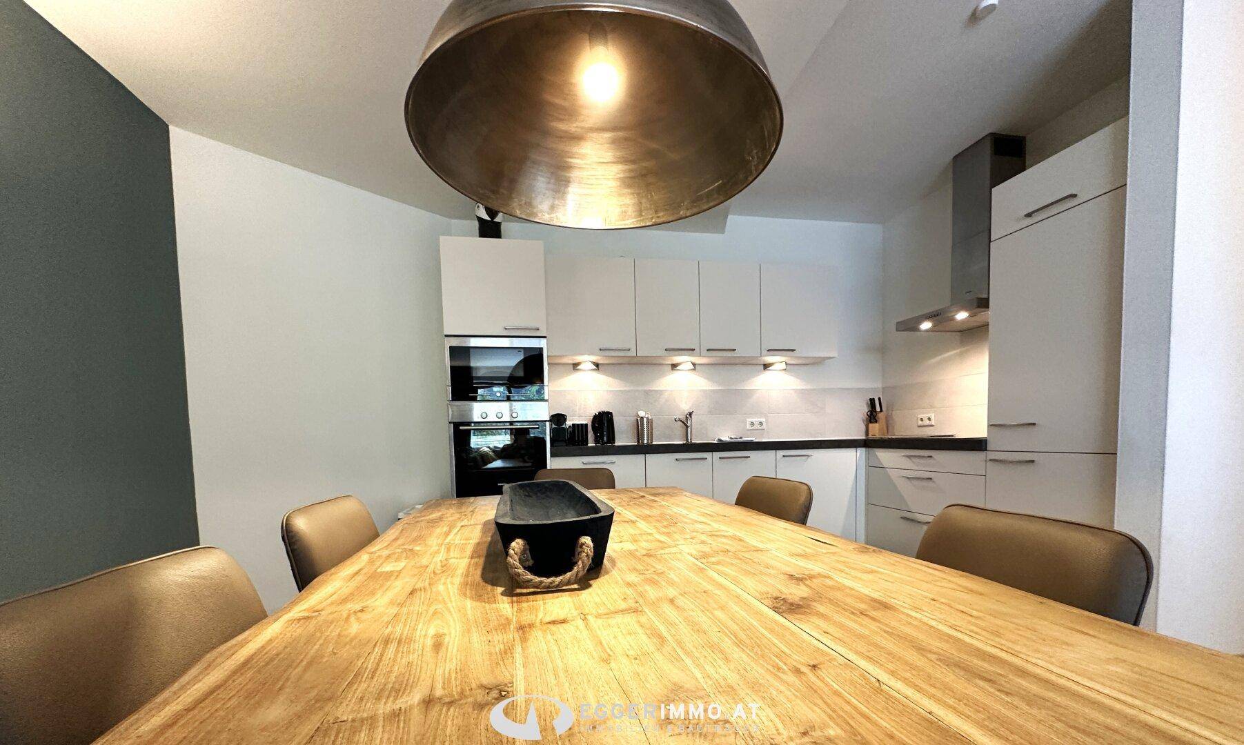 Moderne Einbauküche mit weißen Schränken, dunkler Arbeitsplatte und großem Esstisch unter einer markanten Lampe.