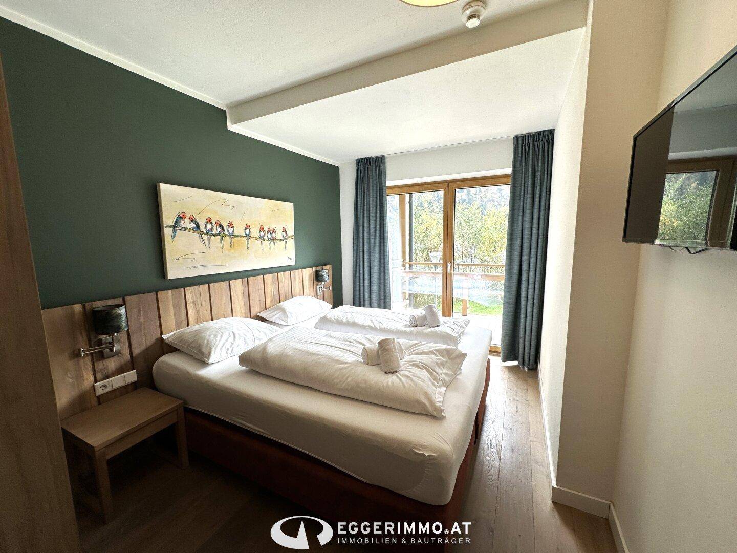 Schlafzimmer mit Doppelbett, Holzkopfteil, grüner Akzentwand und Zugang zum Balkon mit Bergblick.