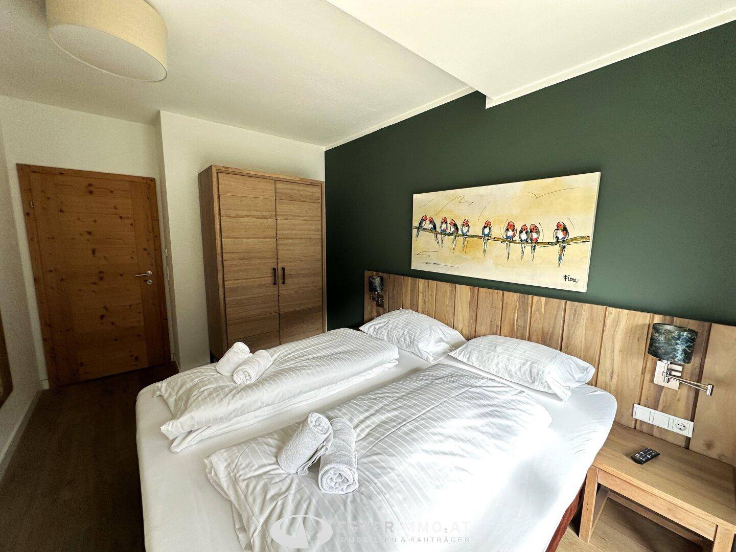 Schlafzimmer mit Doppelbett, Holzschrank und Kunstwerk über dem Bett, das eine gemütliche Atmosphäre schafft.