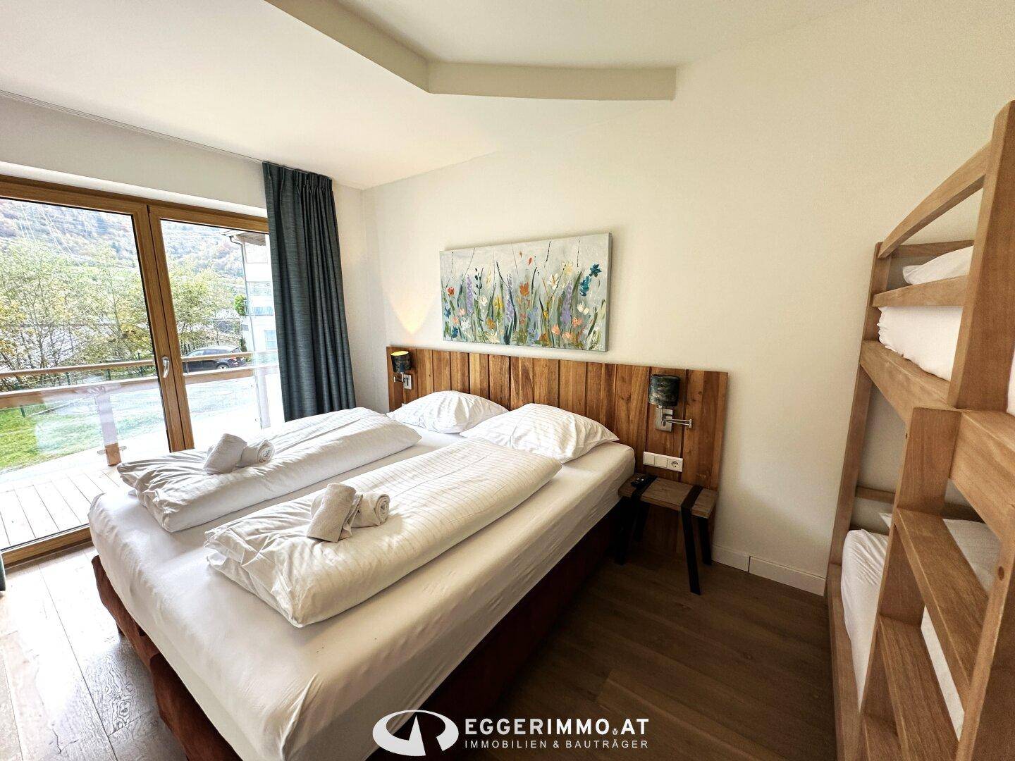 Schlafzimmer mit Doppelbett, Etagenbett und großen Fenstern, die Zugang zu einem Außenbereich bieten.