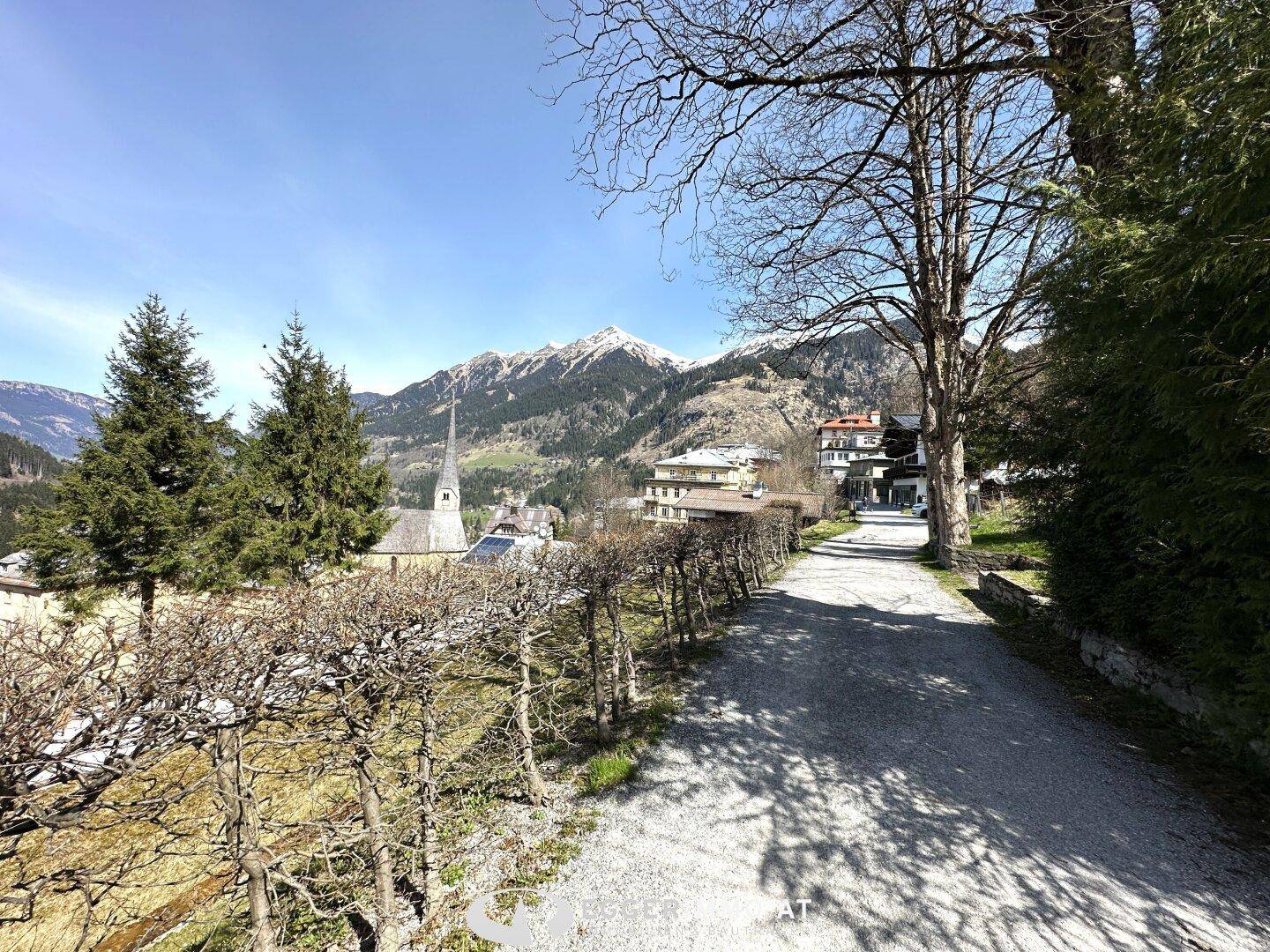 Kiesweg führt durch Bäume mit Blick auf ein Bergdorf und schneebedeckte Berge im Sonnenschein.