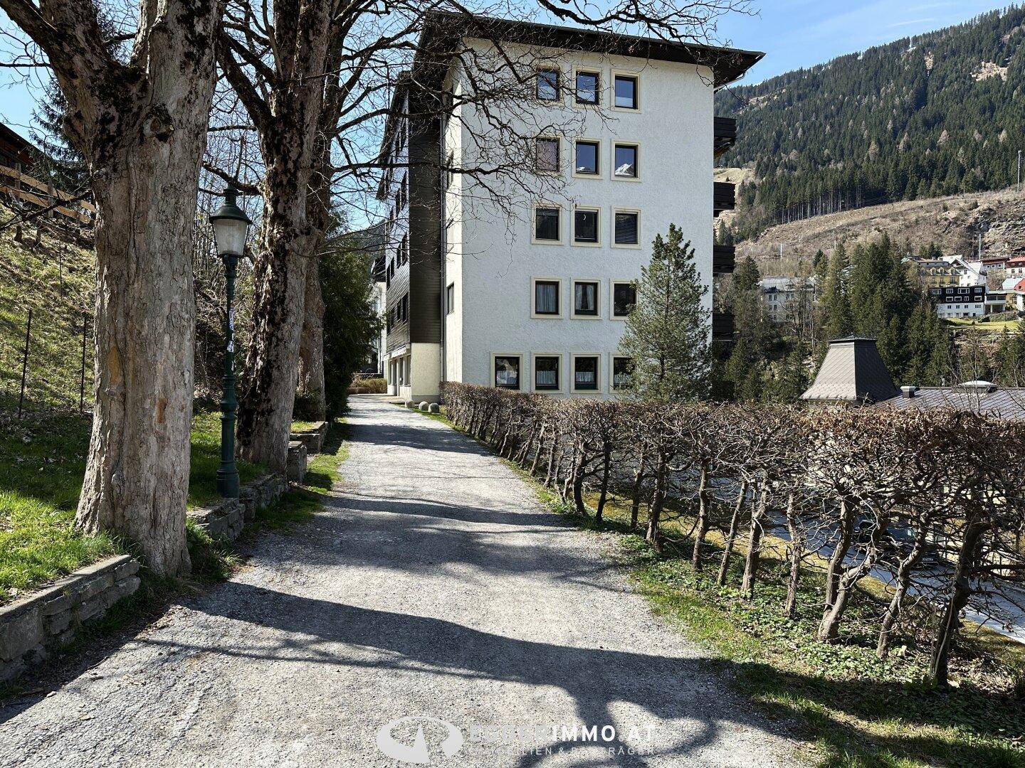 Weiß verputztes Gebäude mit vielen Fenstern, umgeben von Bäumen und einer Hecke.