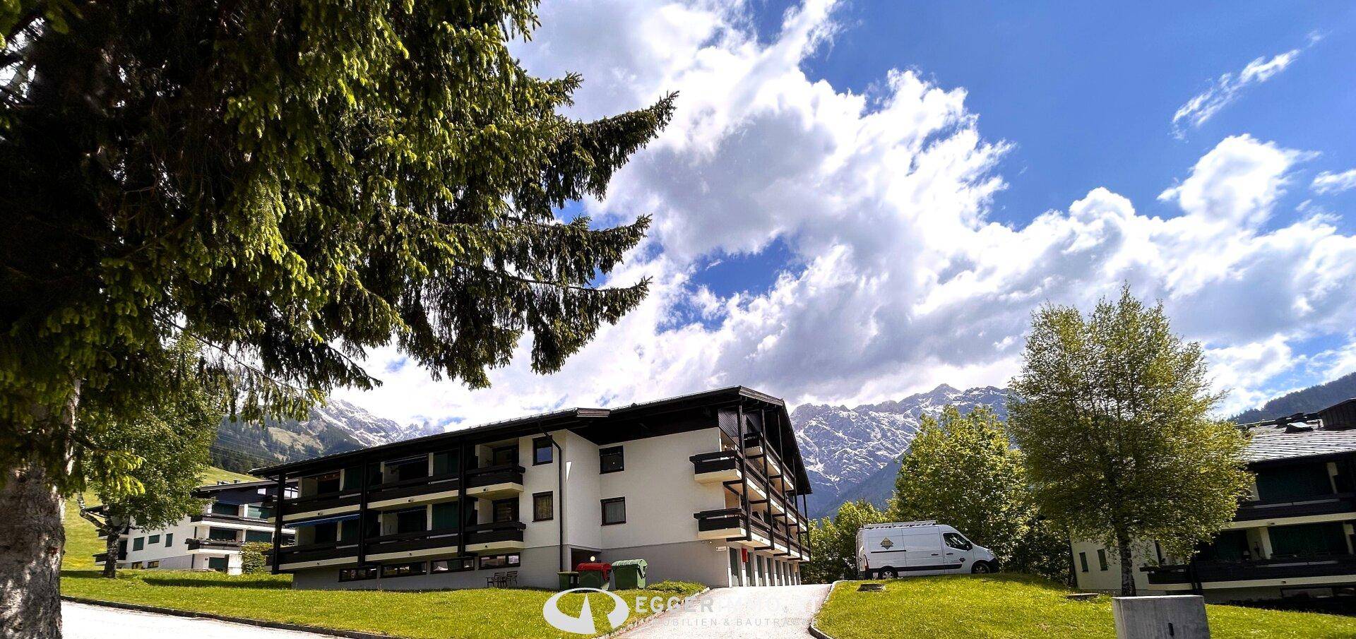 Gebäudekomplex in alpiner Landschaft mit Bäumen und Bergen im Hintergrund unter blauem Himmel.