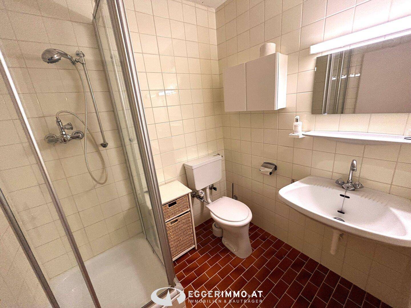 Badezimmer mit Duschkabine, WC, Waschbecken und hellen Fliesen im 80er-Jahre-Stil.