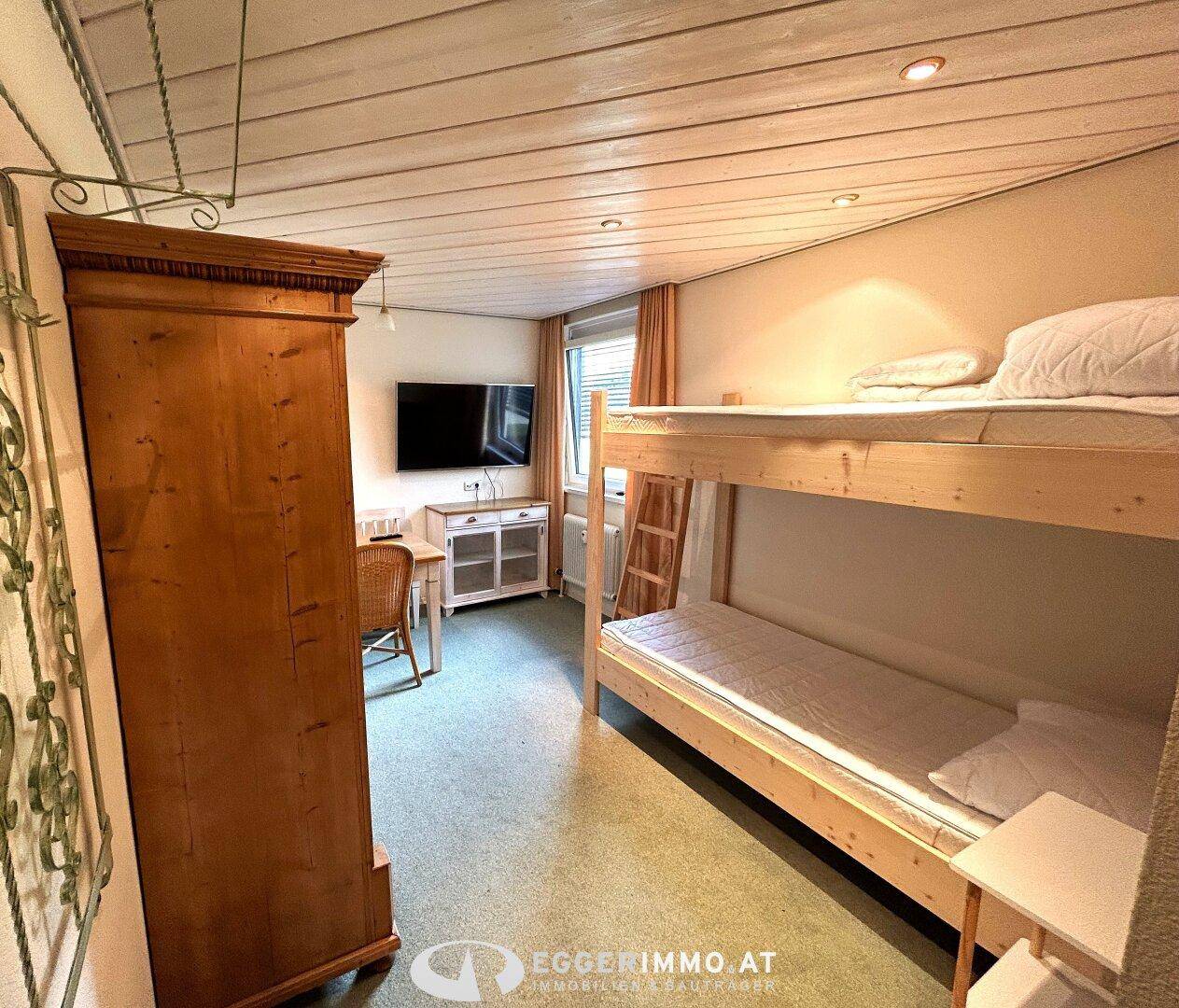 Schlafzimmer mit Etagenbett, Fernseher und traditionellem Holzschrank in warmer Atmosphäre.