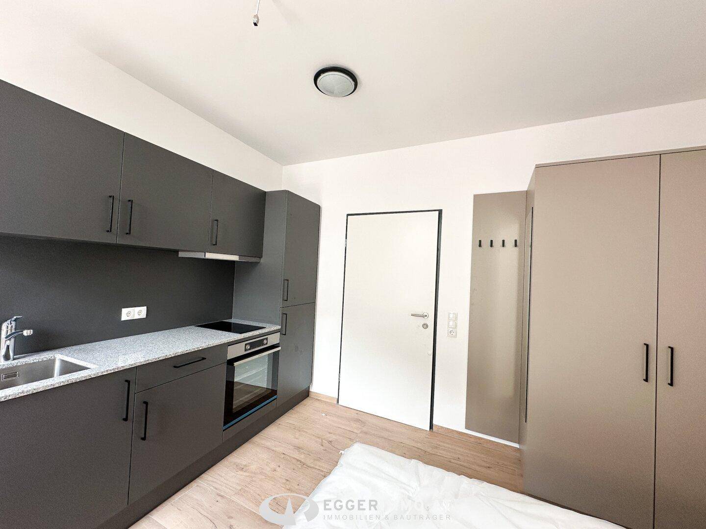 Kompakte Küchenzeile mit grauen Schränken, Kochfeld und Ofen, daneben ein Kleiderschrank.