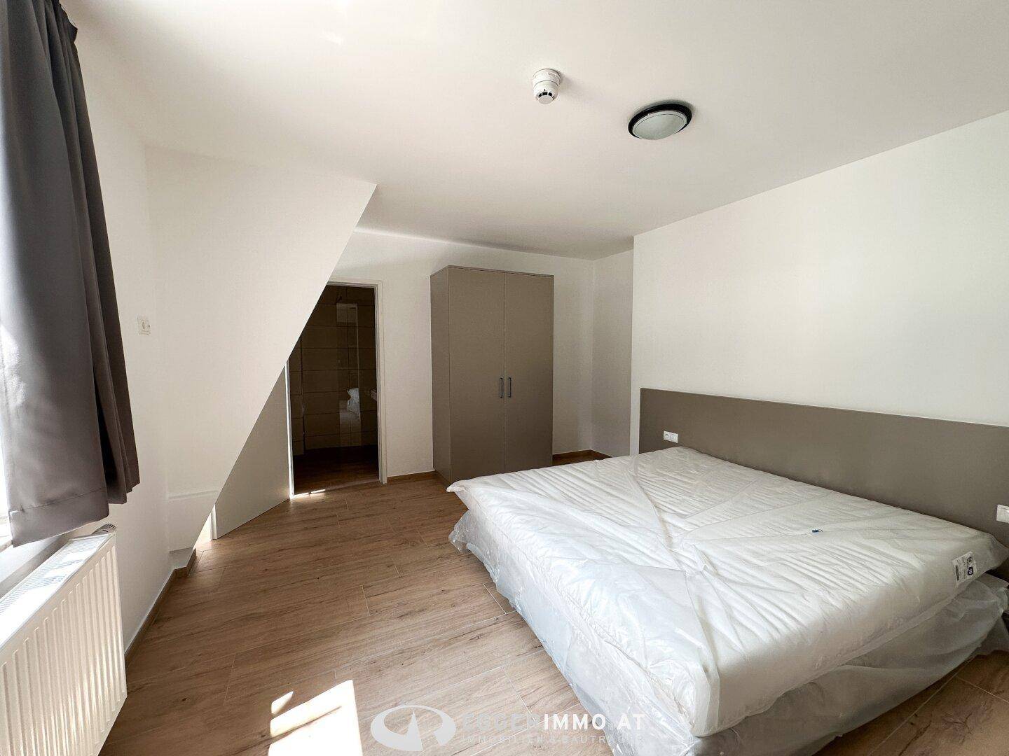 Helles Schlafzimmer mit Holzboden, Kleiderschrank und Zugang zu einem modernen Badezimmer.