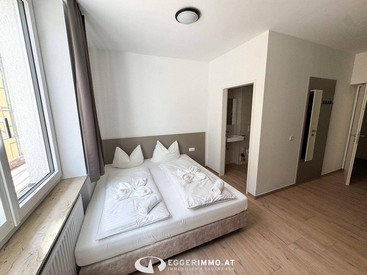 Schlafzimmer mit Doppelbett und direktem Zugang zu einem modernen Badezimmer mit Waschbecken.