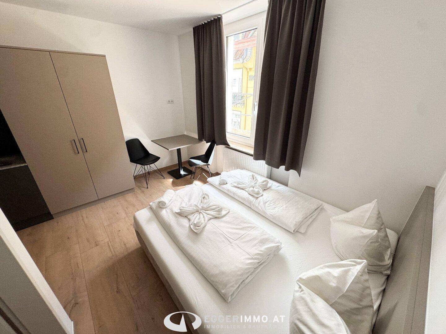 Helles Schlafzimmer mit Doppelbett, Kleiderschrank und Blick nach draußen durch große Fenster.
