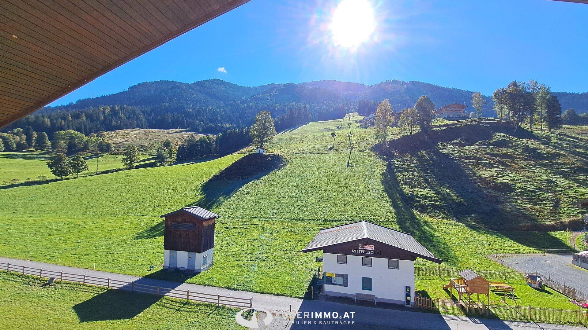 Sonniger Ausblick auf eine grüne Skipiste mit einem kleinen Haus und Spielplatz in der Ferne.