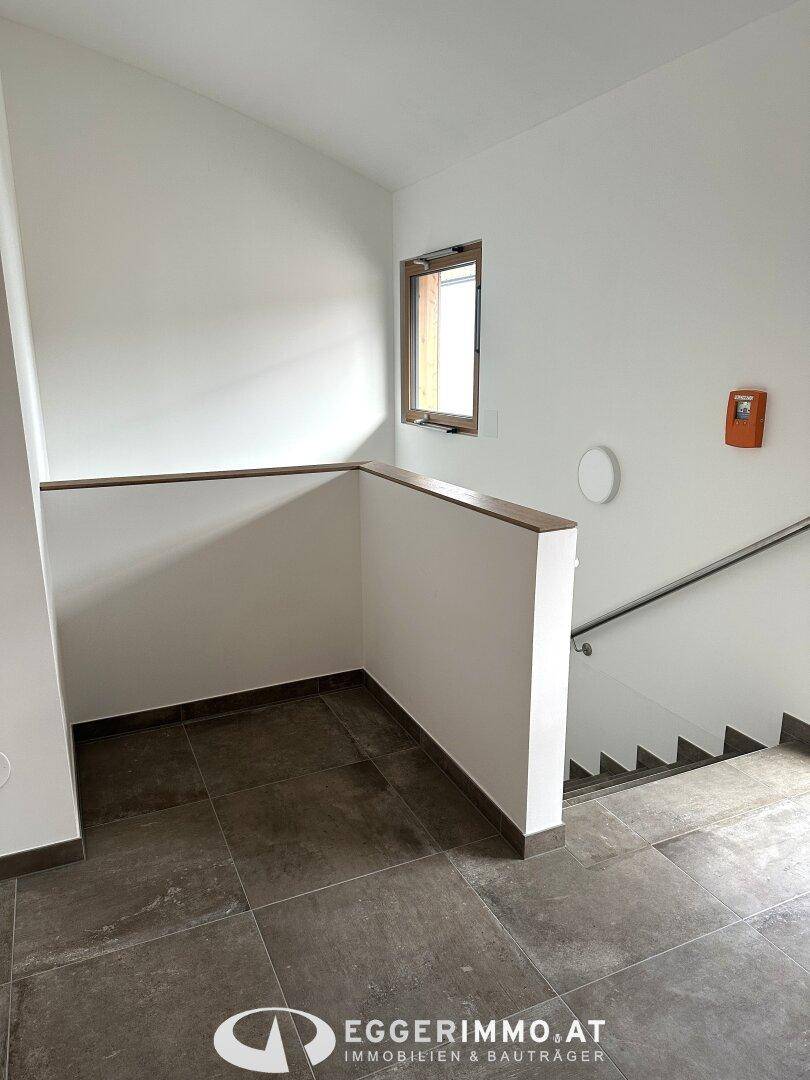 Treppenabsatz mit Fliesenboden, Fenster und schlichtem Geländer in einem modernen Gebäude.