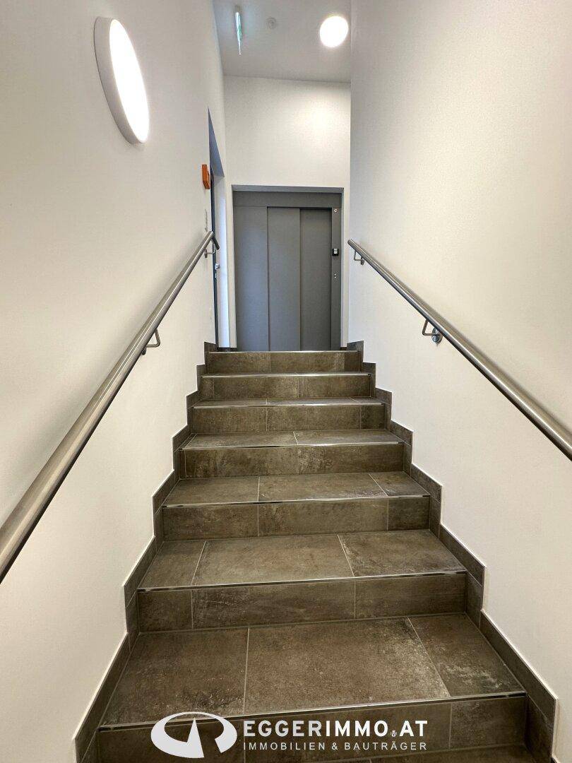 Treppenhaus mit Fliesenboden, Edelstahlgeländer und einem modernen Aufzug im Hintergrund.