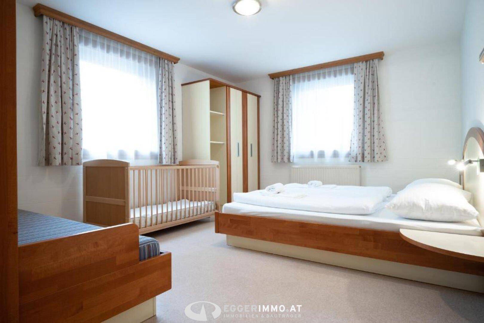 Helles Schlafzimmer mit Doppelbett, Babybett und Kleiderschrank, ausgestattet mit zwei Fenstern.