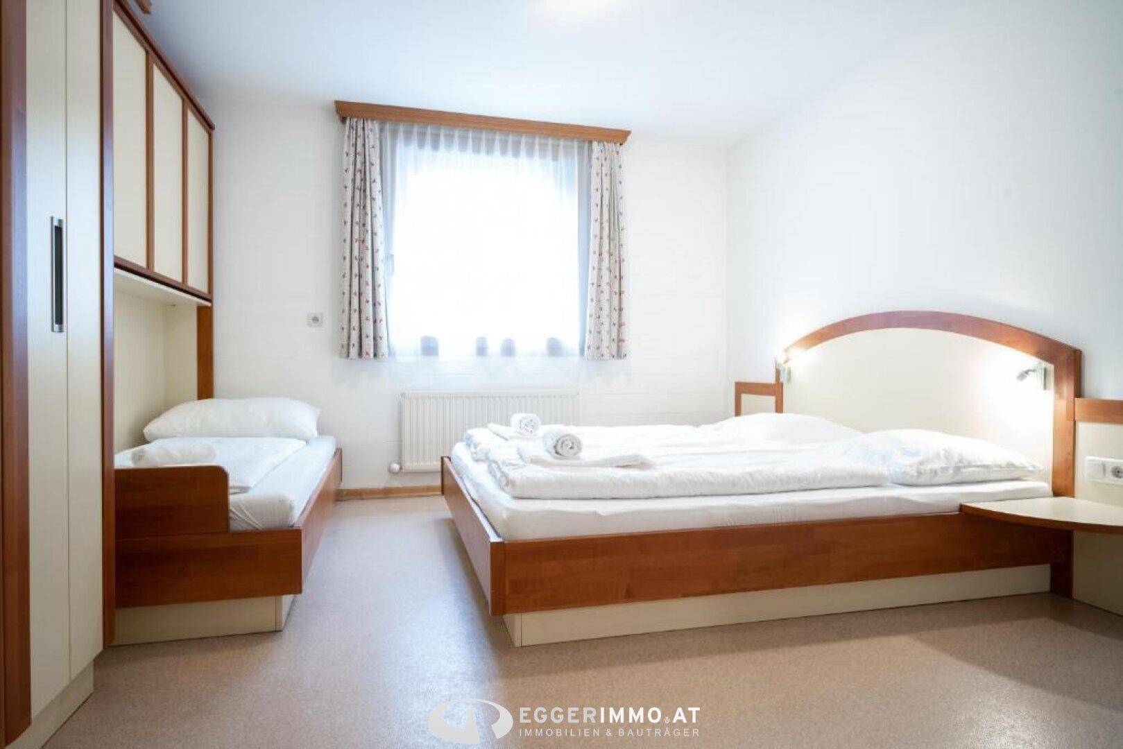 Helles Schlafzimmer mit Doppelbett und Einzelbett, ausgestattet mit Kleiderschrank und Fenster.