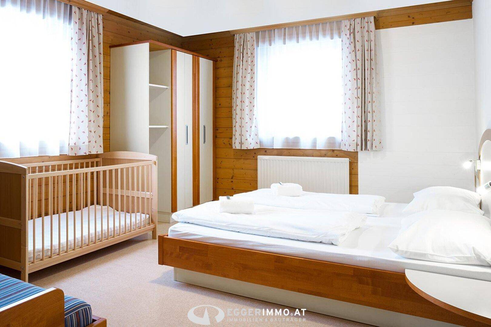 Gemütliches Schlafzimmer mit Holzvertäfelung, Doppelbett, Babybett und Kleiderschrank, hell durch Fenster.