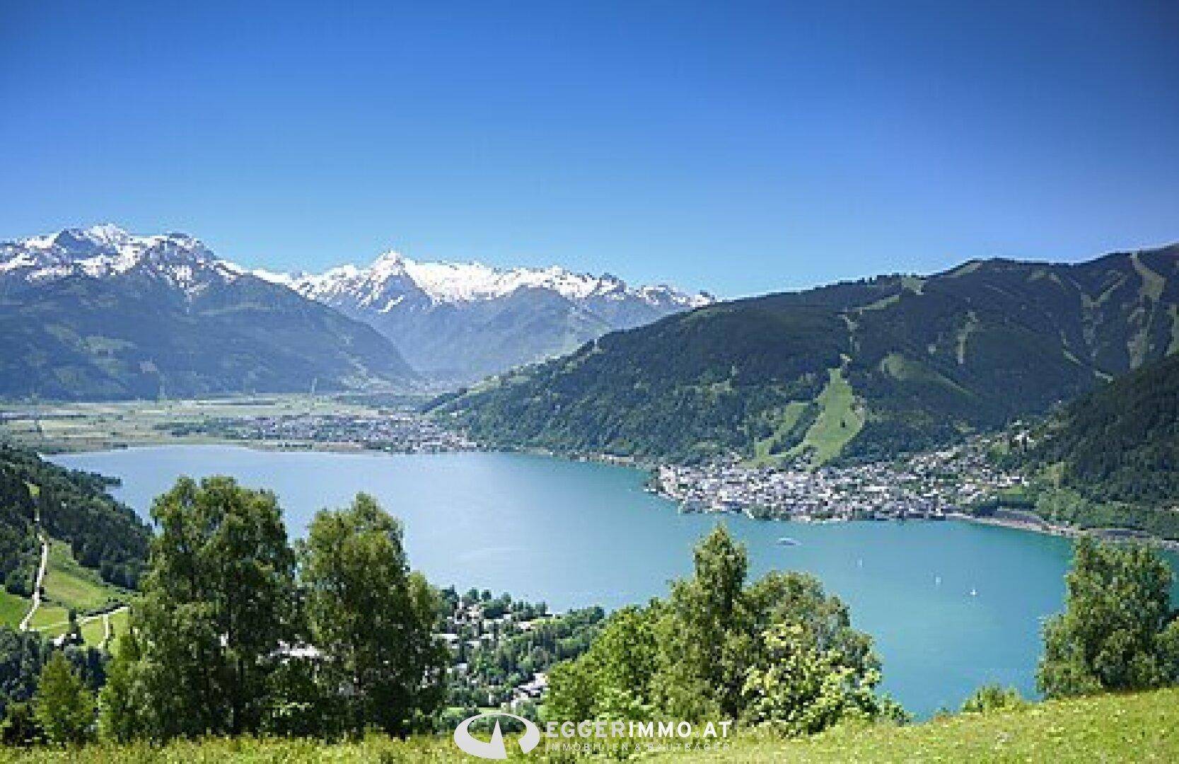 Weitläufiger Blick auf einen See und schneebedeckte Berge, umgeben von grüner Landschaft.