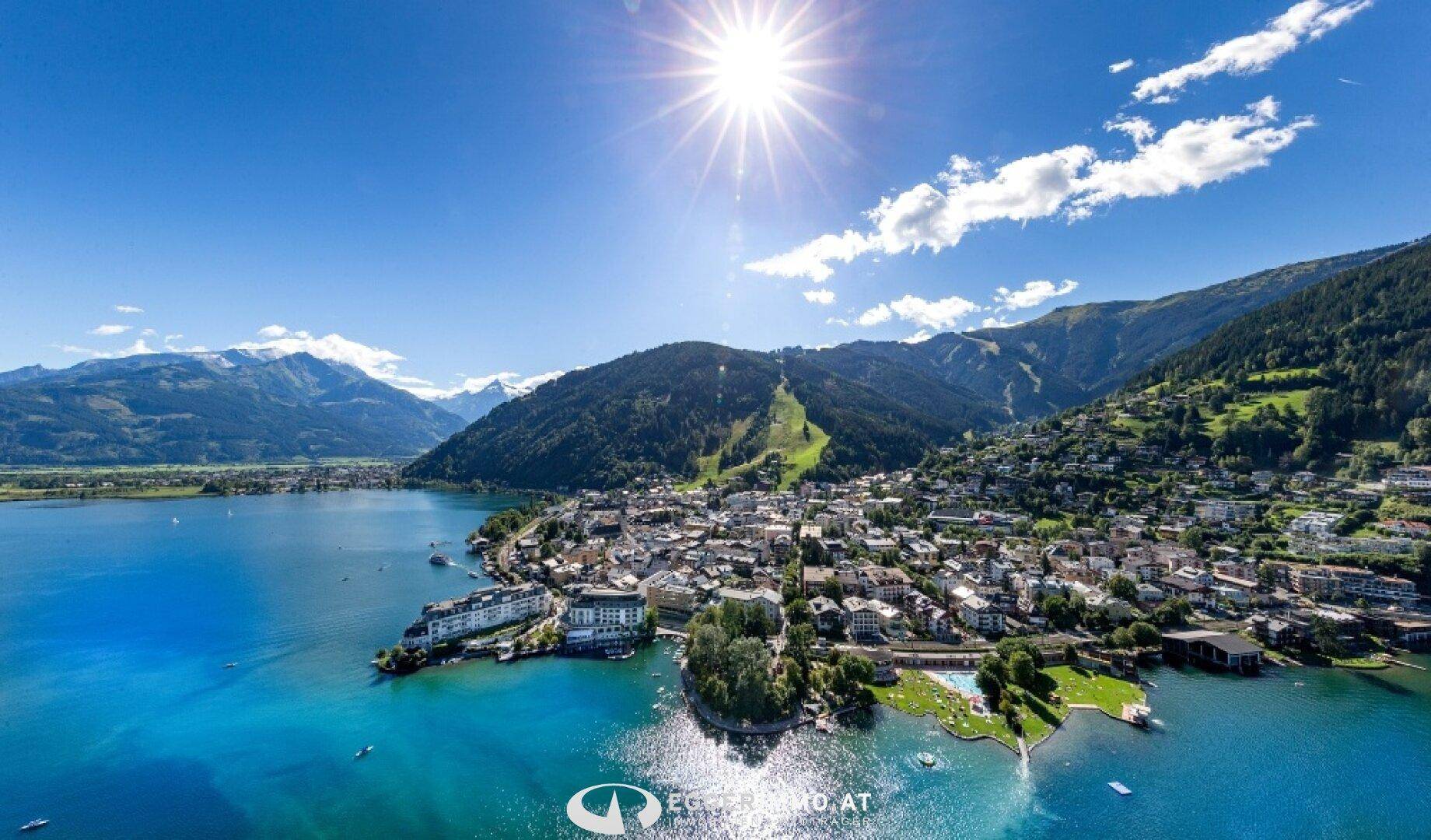 Sommerlicher Panoramablick auf eine Stadt am See, umgeben von grünen Bergen unter blauem Himmel.