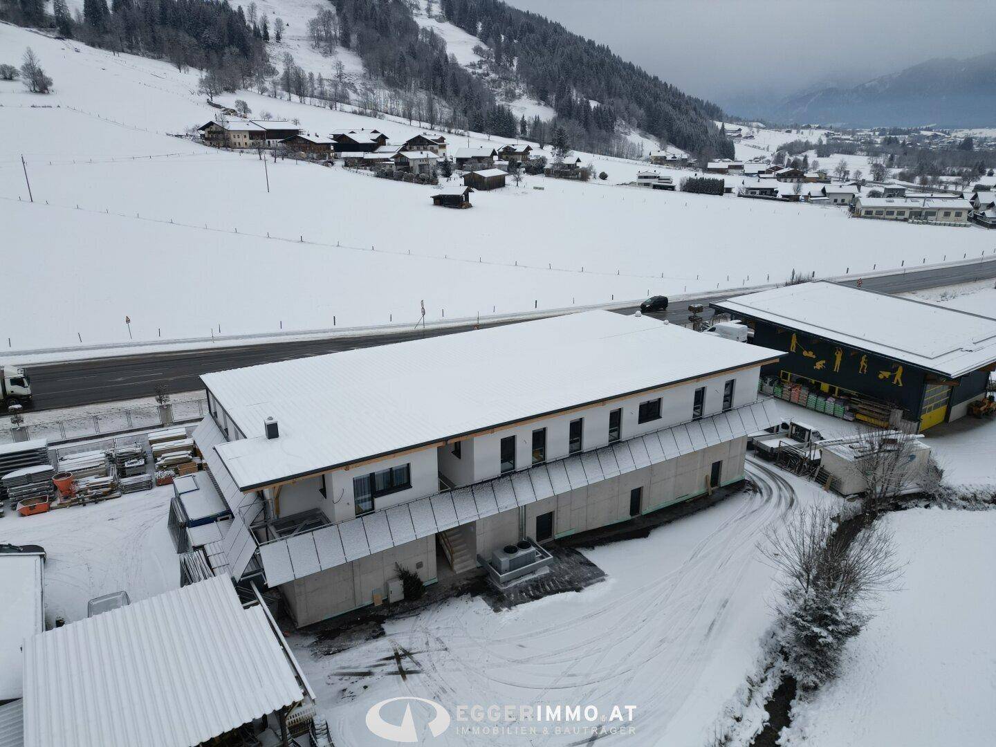 Luftaufnahme eines modernen Gewerbegebäudes mit schneebedecktem Dach und umliegender Winterlandschaft mit Straße.