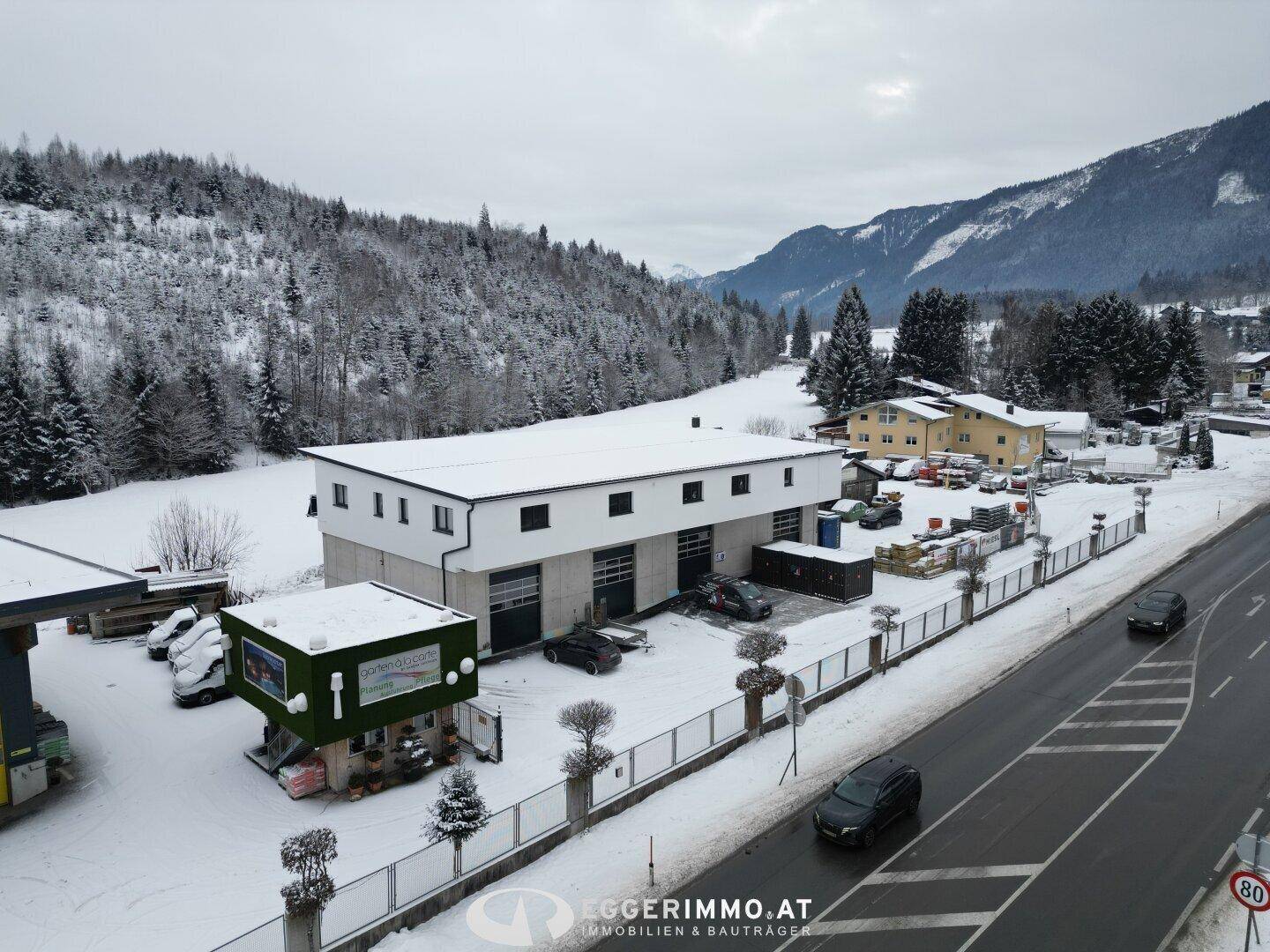 Gewerbeimmobilie an einer Straße gelegen, umgeben von verschneiten Feldern und Bergen unter bewölktem Himmel.