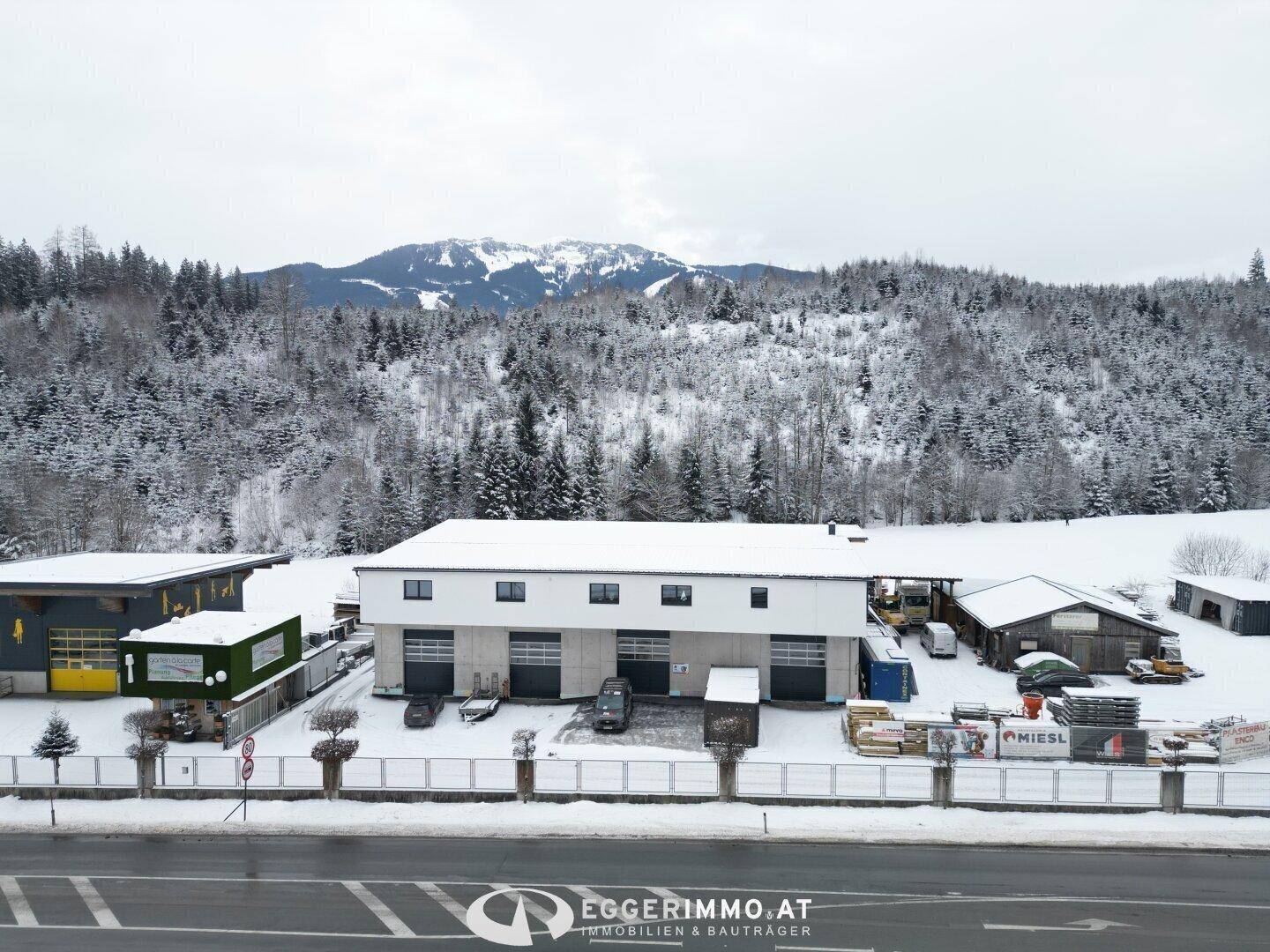 Vorderansicht des Gewerbegebäudes im Winter, mit verschneiten Bäumen und einem beeindruckenden Bergpanorama im Hintergrund.