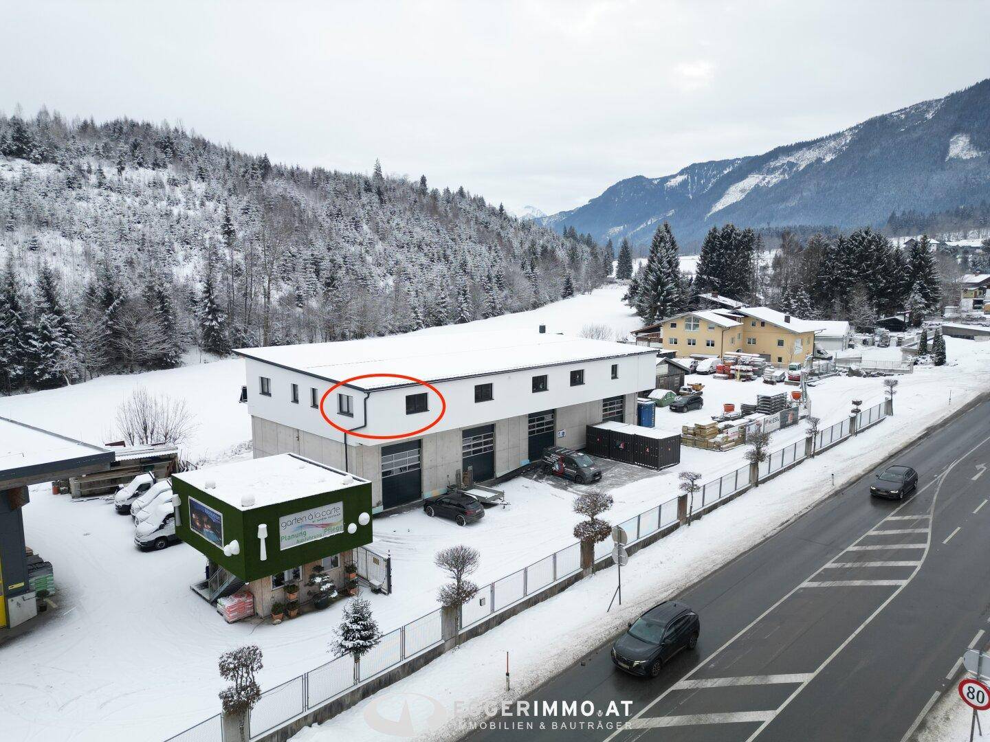 Luftaufnahme eines modernen Gewerbegebäudes im Winter, umgeben von verschneiten Bergen und Wäldern, mit einer Straße im Vordergrund.