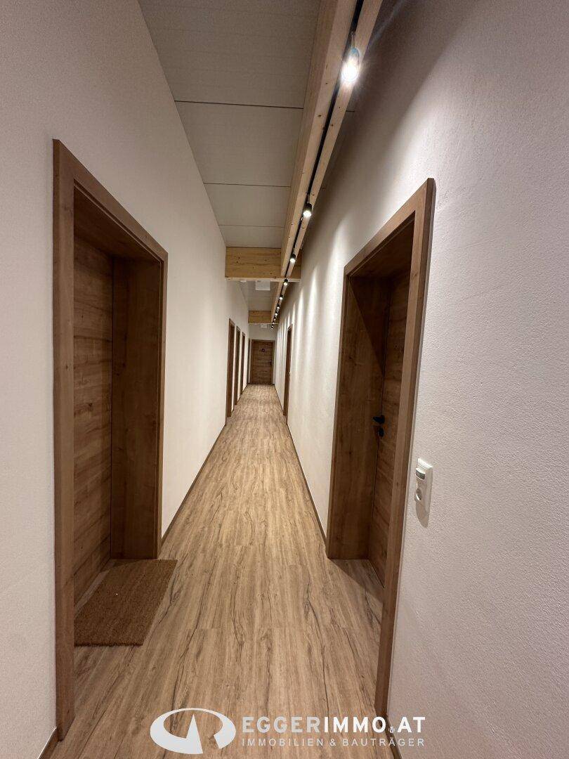 Langer, heller Flur mit Holztüren und modernem Vinylboden, beleuchtet von Deckenspots und sichtbaren Holzbalken.
