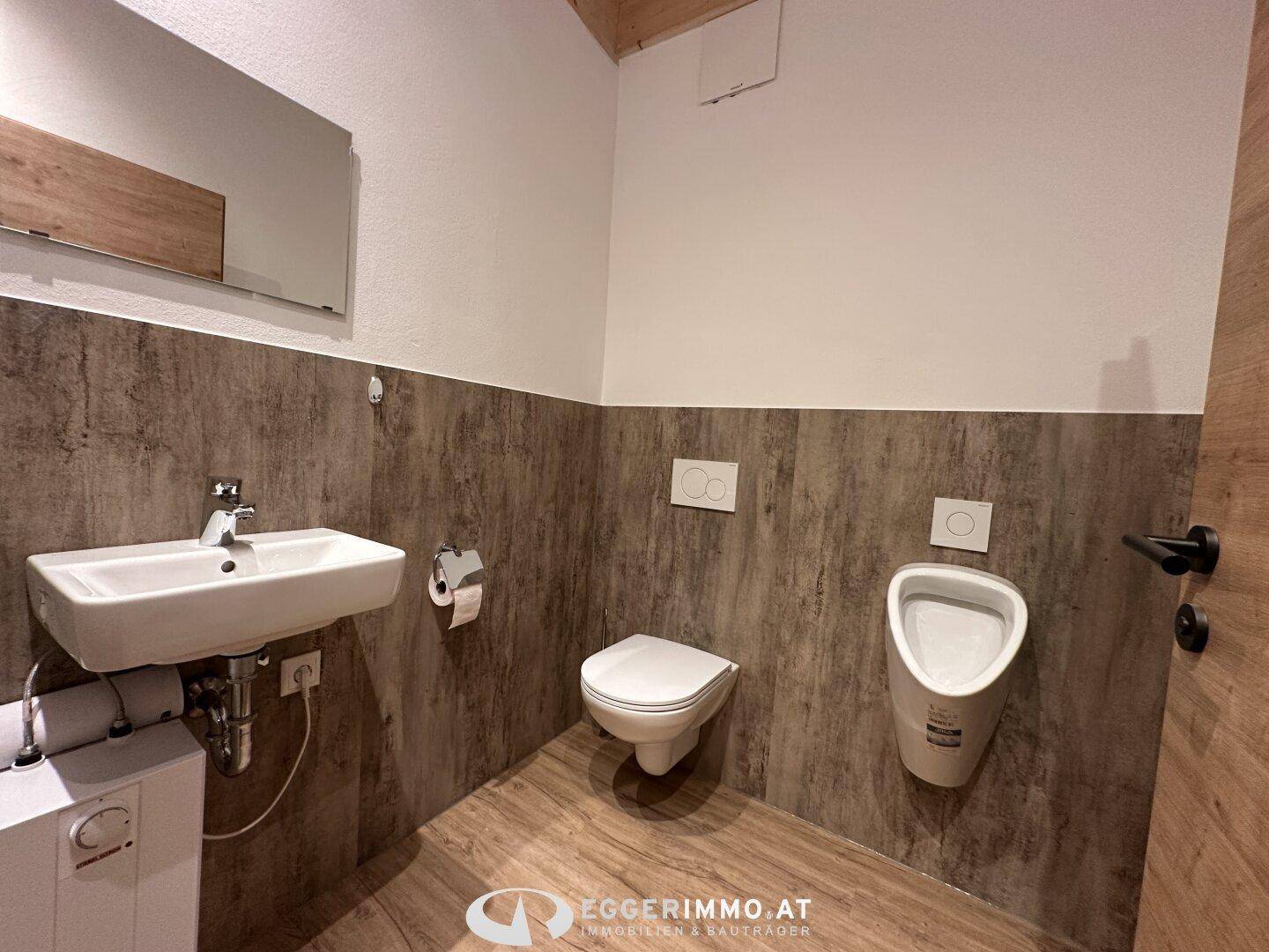 Modernes WC mit Wand-WC, Urinal, Waschbecken und Holzoptik-Fliesen an Wand und Boden.