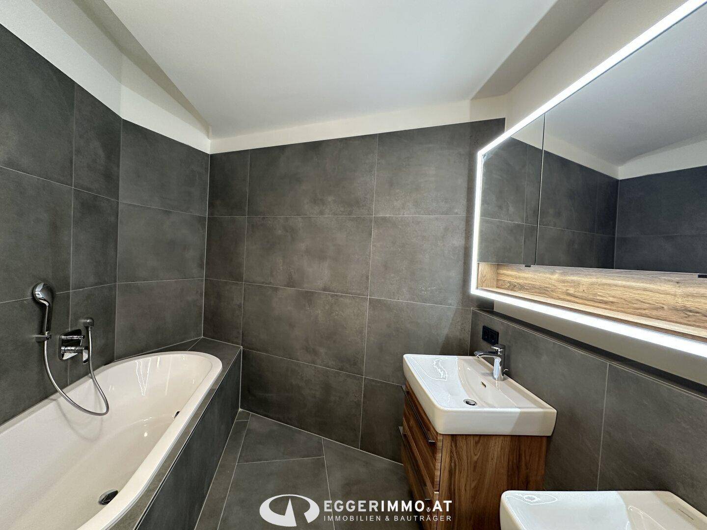 Modernes Badezimmer mit dunklen Fliesen, Badewanne und stilvollem Waschtisch mit beleuchtetem Spiegel.