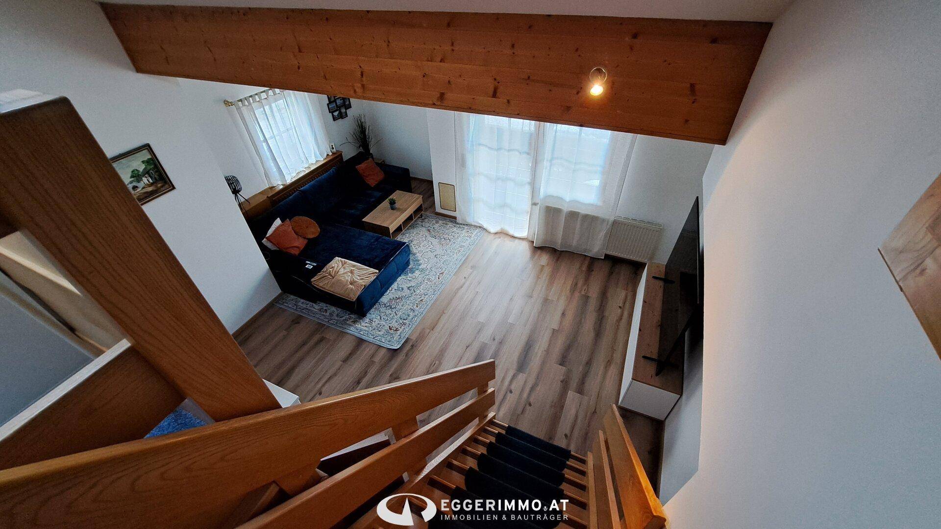 Blick von der Holztreppe in ein helles Wohnzimmer mit Holzboden und blauer Couch.