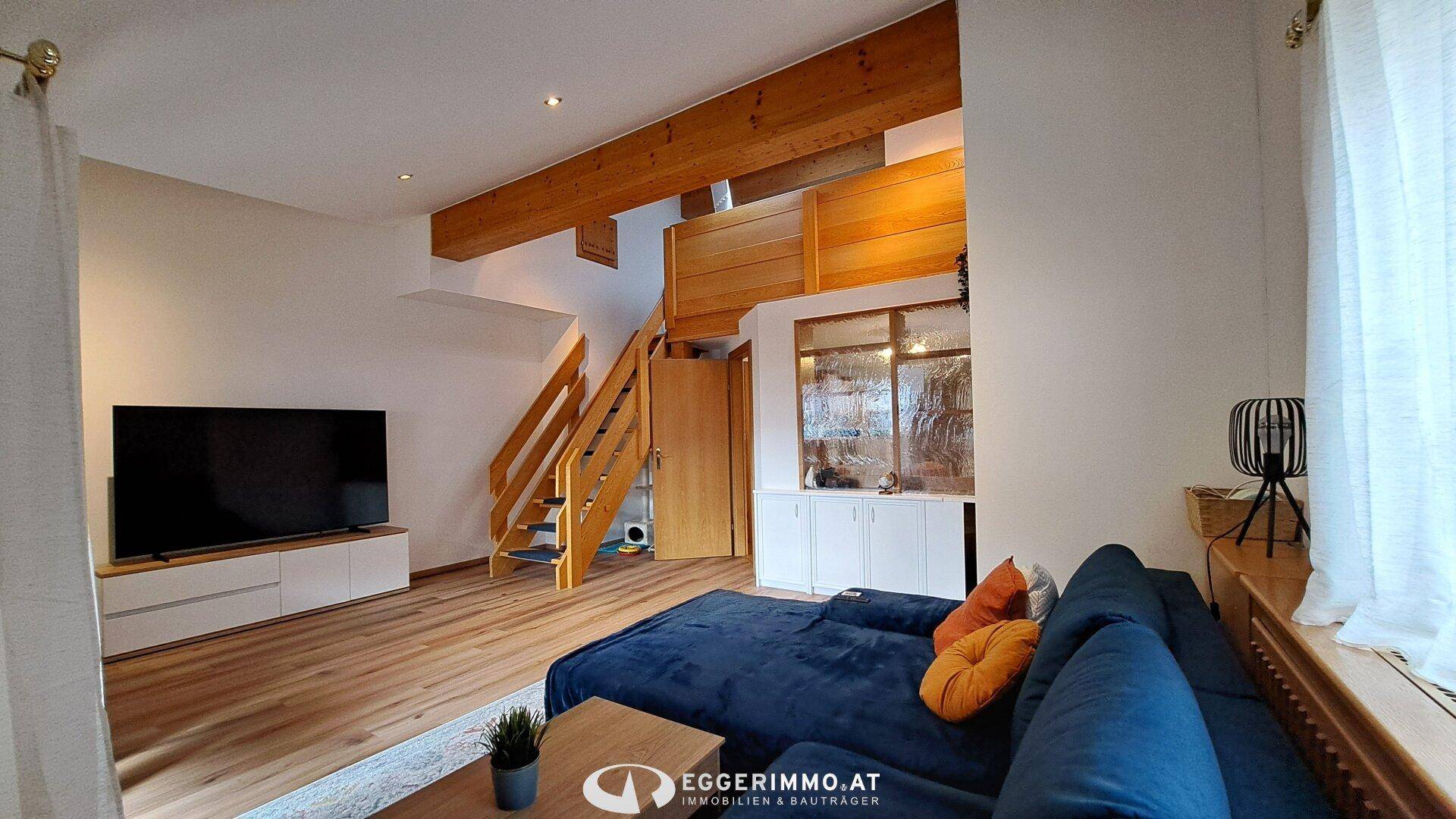 Modernes Wohnzimmer mit Holzbalken, blauer Couch und großem Fernseher, hell und offen.