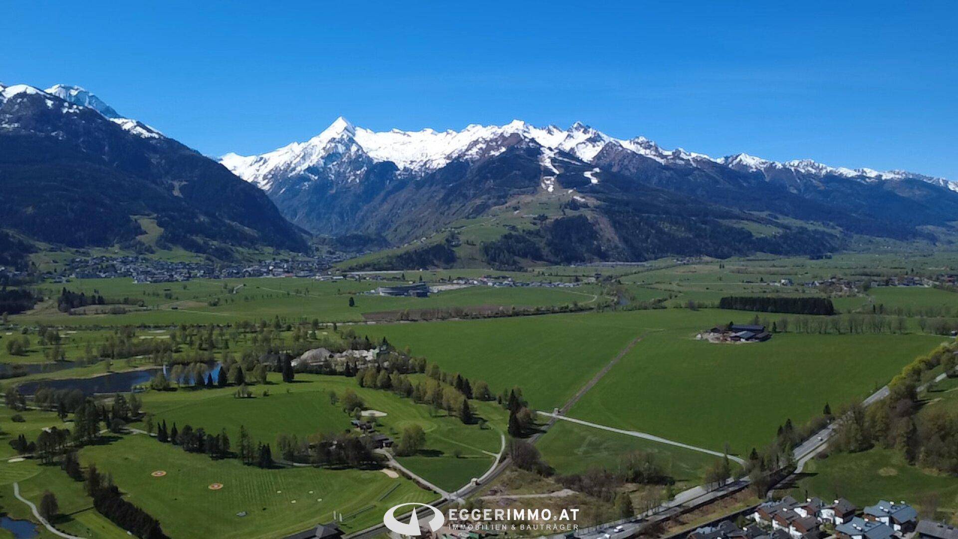 Weitläufige Landschaft mit schneebedeckten Bergen und grünen Feldern unter blauem Himmel.