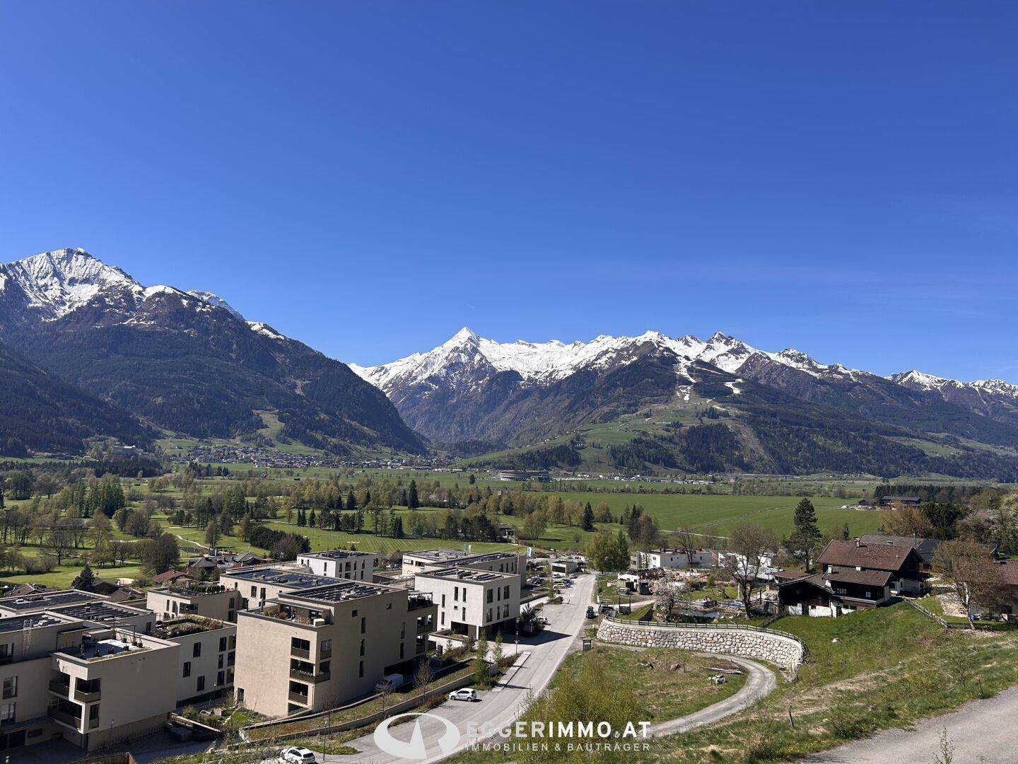 Panoramablick auf die schneebedeckten Berge, grüne Täler und eine Wohnsiedlung.