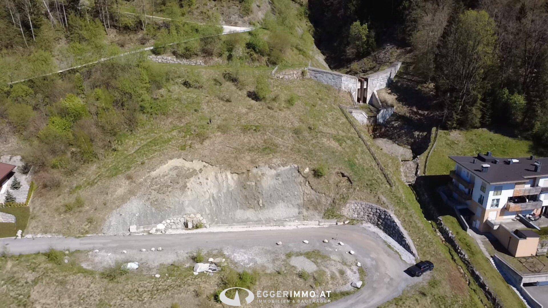 Luftaufnahme eines Hanggrundstücks mit angrenzendem Gebäude und einer Zufahrtsstraße.