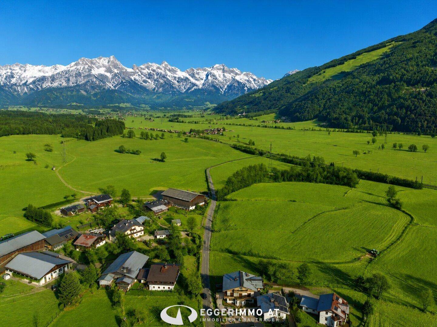 Panoramische Luftaufnahme eines weiten Tals mit verstreuten Häusern, grünen Feldern und majestätischen Bergen.