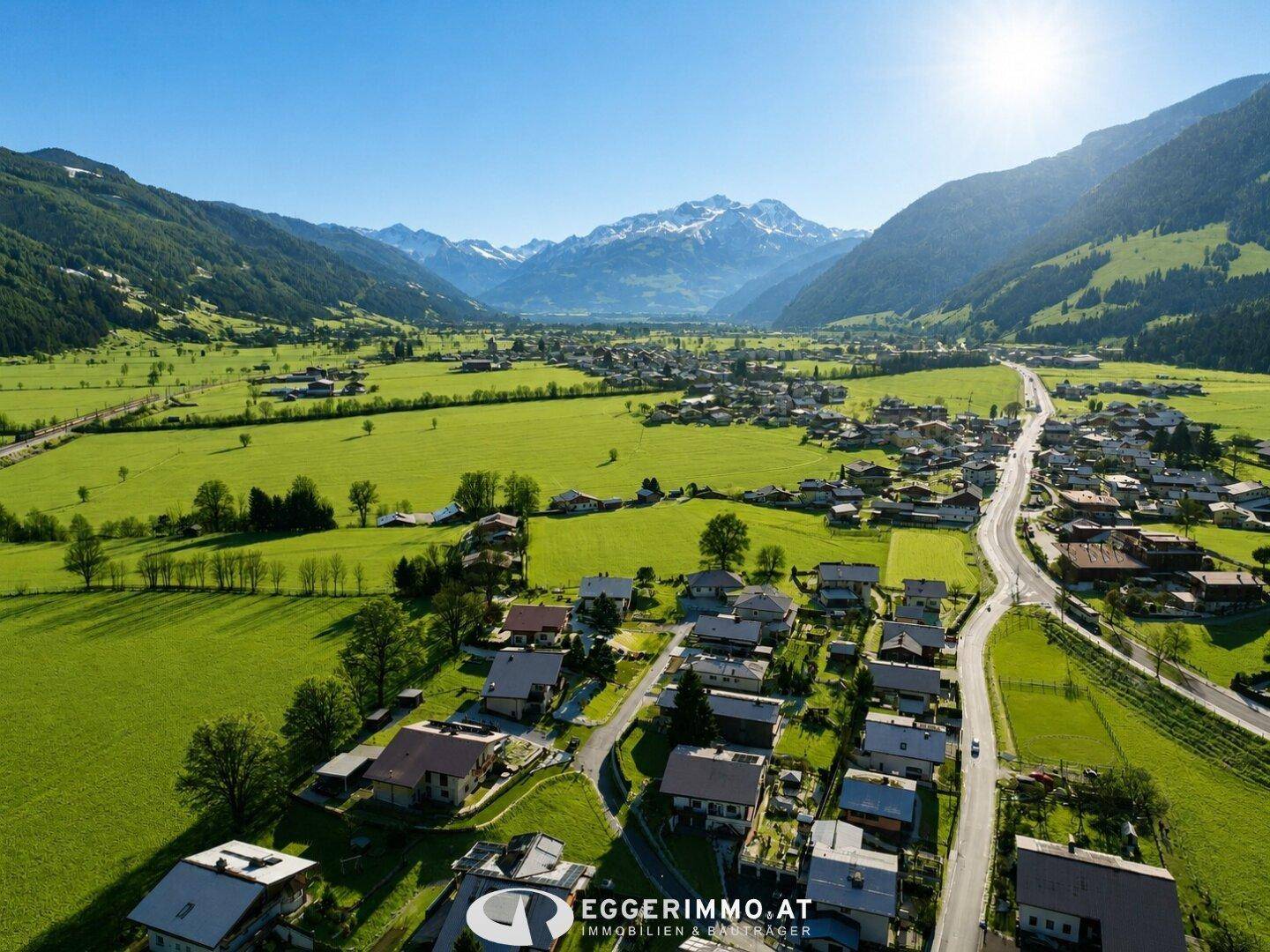 Weitläufige Luftaufnahme eines sonnigen Tals mit Dorf, grünen Wiesen und schneebedeckten Bergen im Hintergrund.