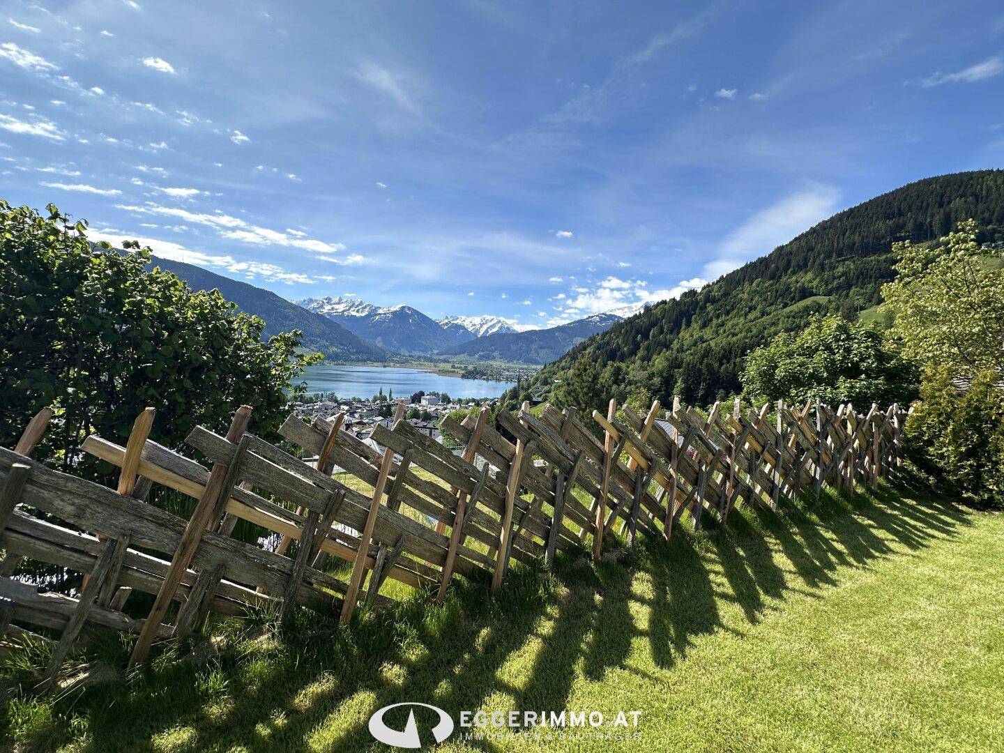 Malerischer Blick auf einen See und schneebedeckte Berge von einem grünen Gartenbereich mit Holzzaun.