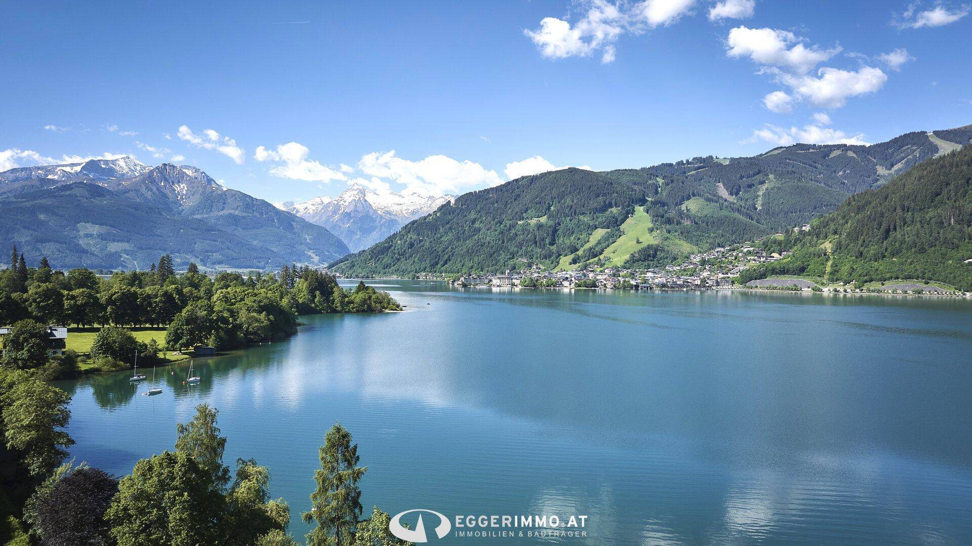 Weitläufiger Blick über den glitzernden See mit schneebedeckten Berggipfeln und grünen Ufern unter blauem Himmel.