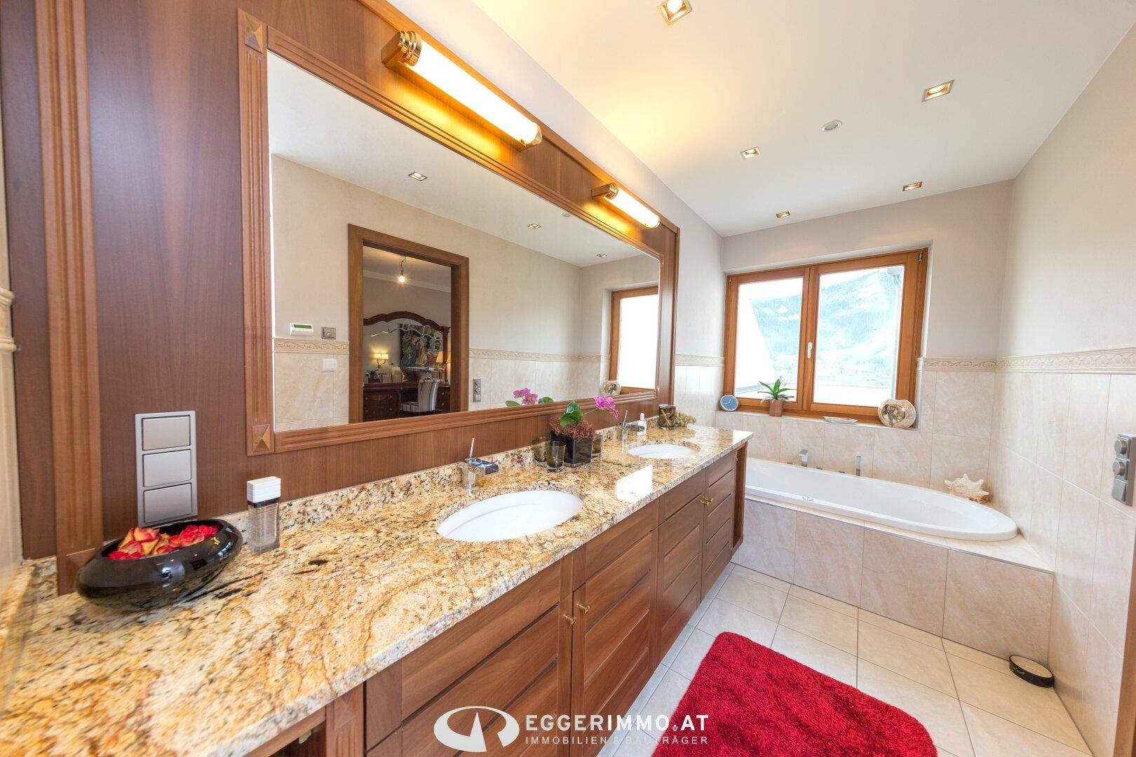 Luxuriöses Badezimmer mit Doppelwaschbecken, Granitplatte, großem Spiegel und Badewanne unter dem Fenster.