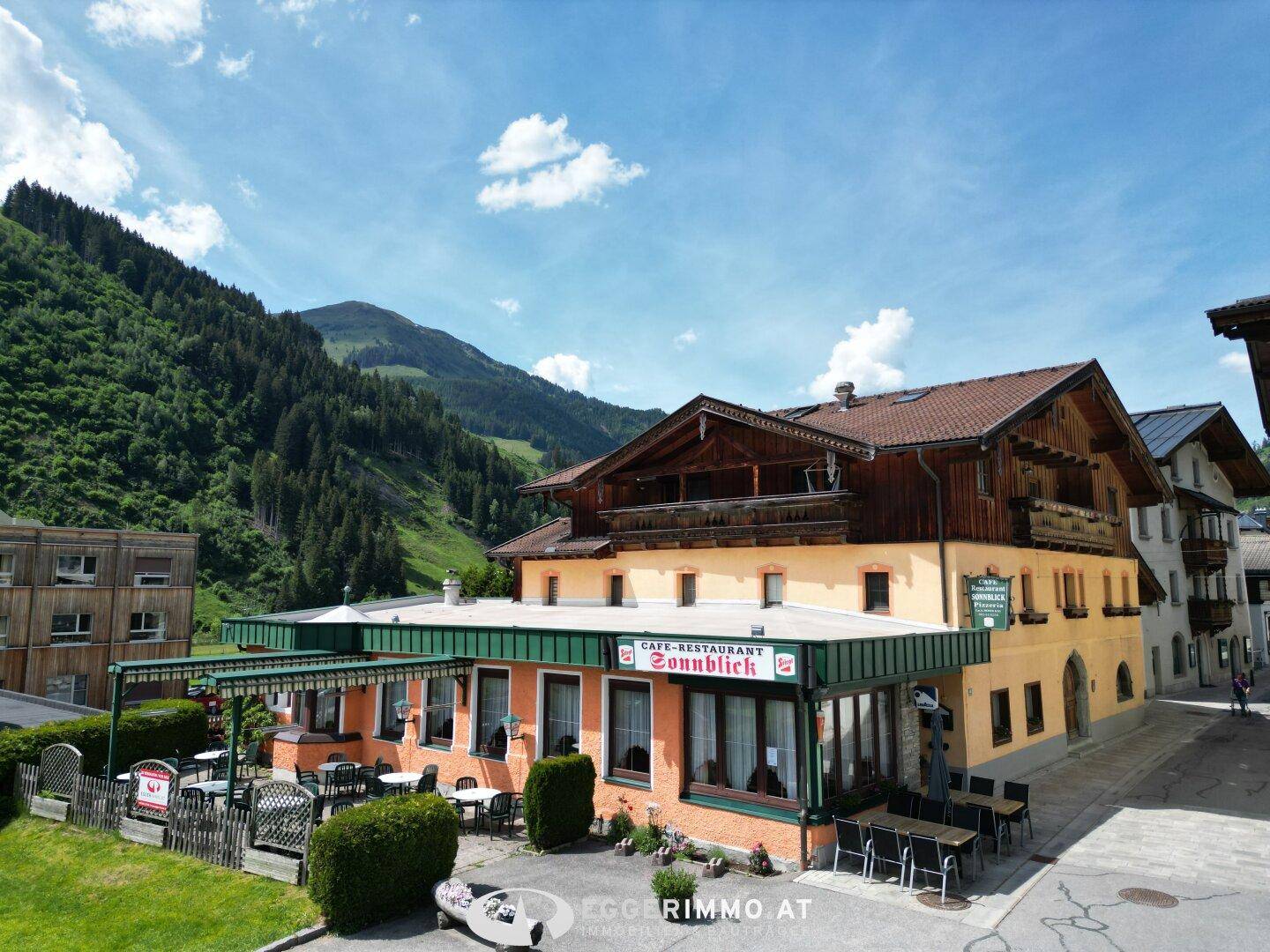 Das Gebäude mit Restaurant und Außenterrasse, umgeben von grünen Bergen unter blauem Himmel.